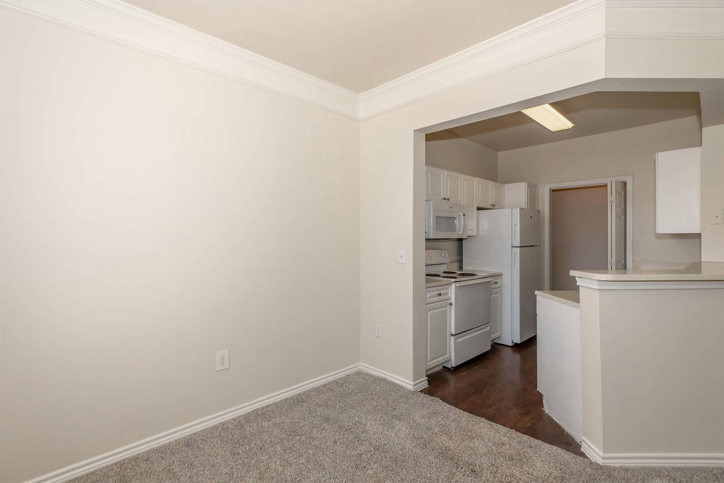 Interior view of a spacious kitchen and living area in an apartment. The kitchen features white cabinets, appliances including a refrigerator, and an open layout leading into the living area with beige walls and carpet flooring. Natural light brightens the space.