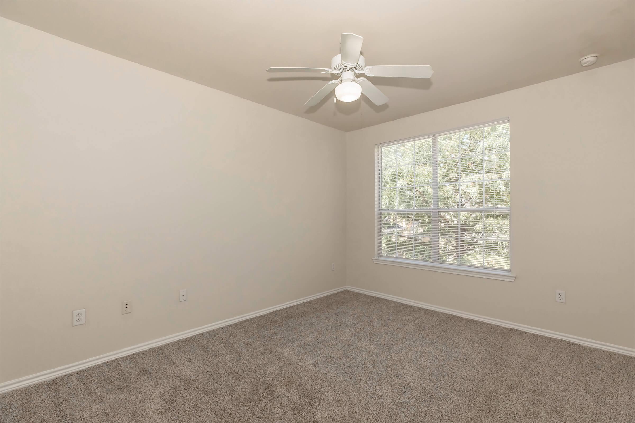 Empty room with beige walls and light brown carpet. A ceiling fan with a light fixture hangs from the ceiling. There is a large window on one wall, allowing natural light to enter, with a view of greenery outside. The space appears clean and ready for furnishing.