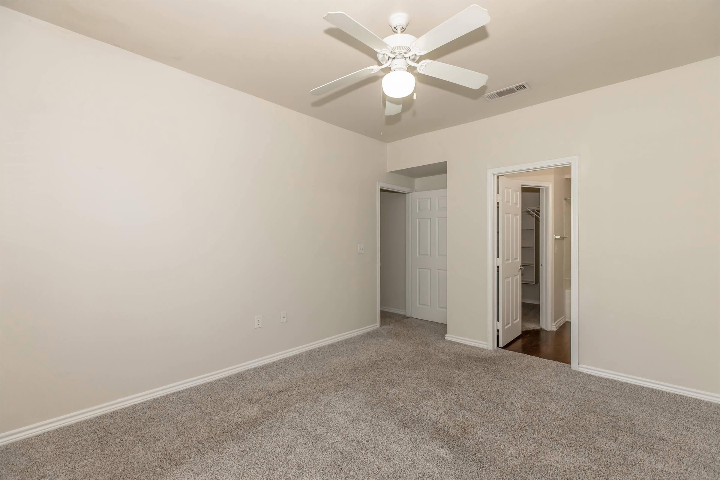 A spacious, empty room with light-colored walls and carpet. It features a ceiling fan and doorways leading to another room and a closet. The overall atmosphere is bright and clean, suitable for various decorative styles.