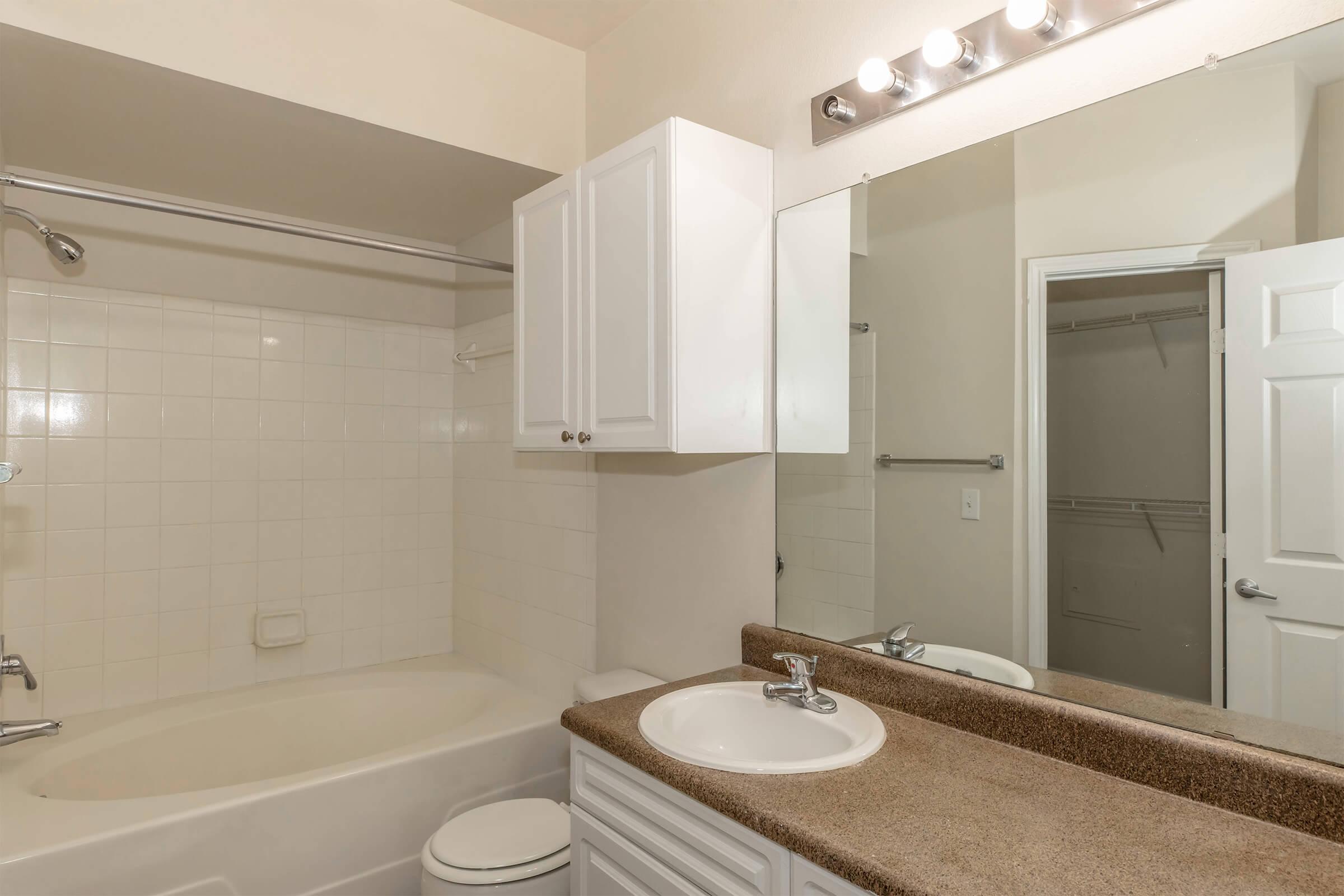 A clean bathroom featuring a white bathtub with a shower, beige walls, and a large mirror above a countertop with a sink. There are white cabinets for storage, and a closet door is visible in the background. The lighting is provided by a row of bulbs above the mirror.