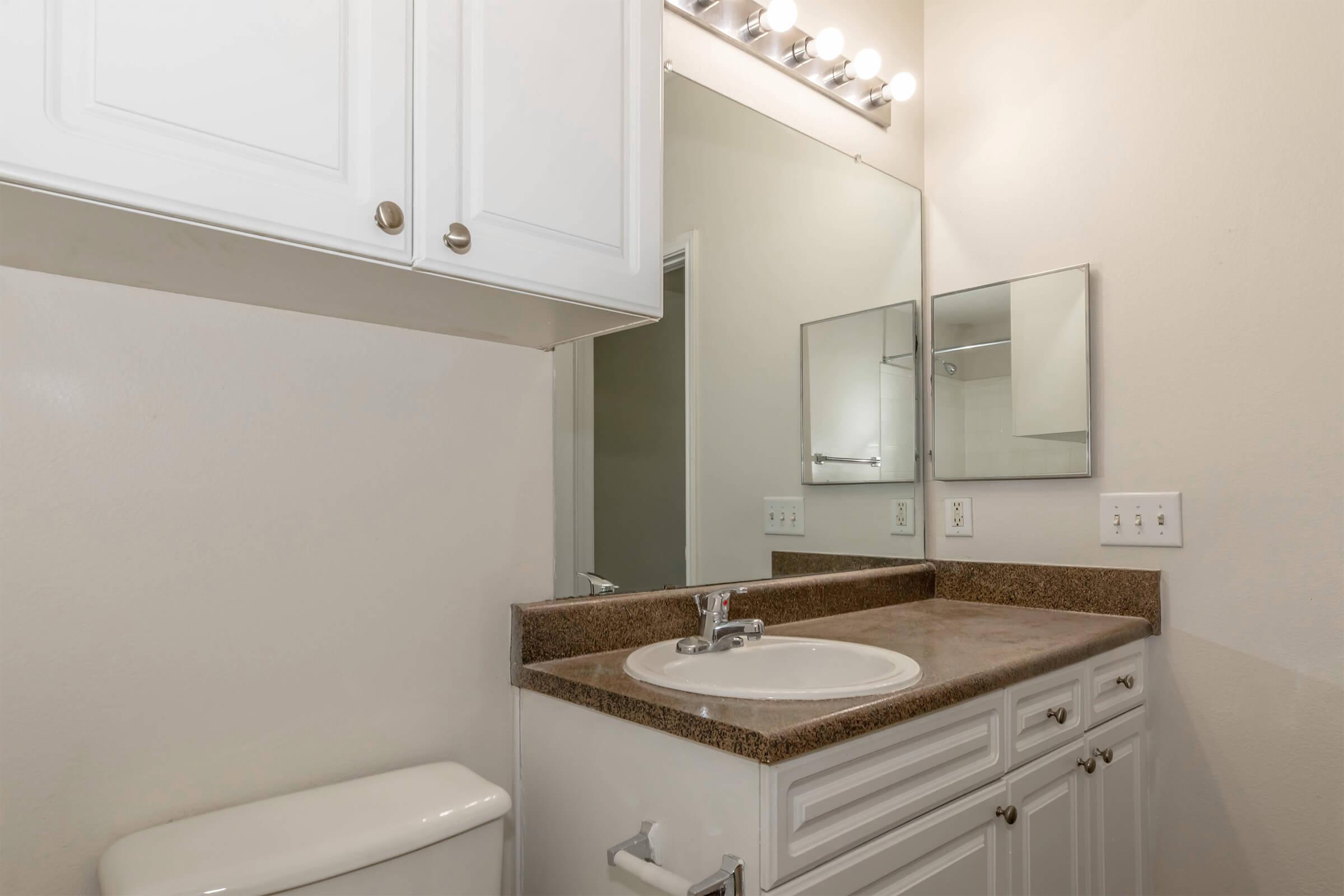 A bright and clean bathroom featuring a white cabinet with silver knobs, a round sink on a brown countertop, and a large mirror. Above the mirror, there are five light bulbs mounted. A white toilet is visible in the corner, and the walls are painted in a light color, contributing to a spacious feel.