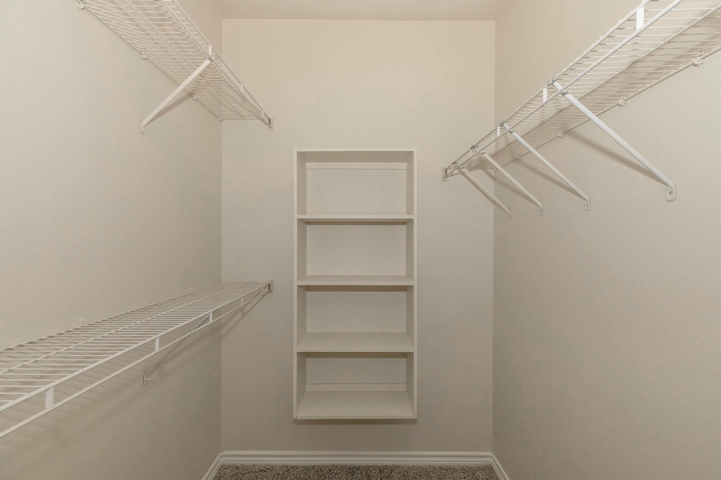 Empty closet storage space featuring white wire shelving on both sides, an open shelf unit in the center, and beige walls. The flooring appears to be carpeted.