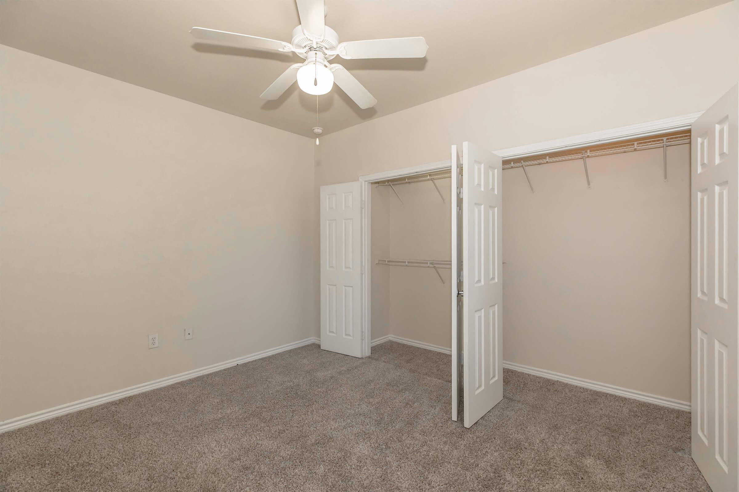 A spacious bedroom with light beige walls and plush carpet flooring. The room features a ceiling fan with a light fixture and two open doors leading to a closet with shelves. Natural light can potentially enter through the room's windows, enhancing the airy feel.