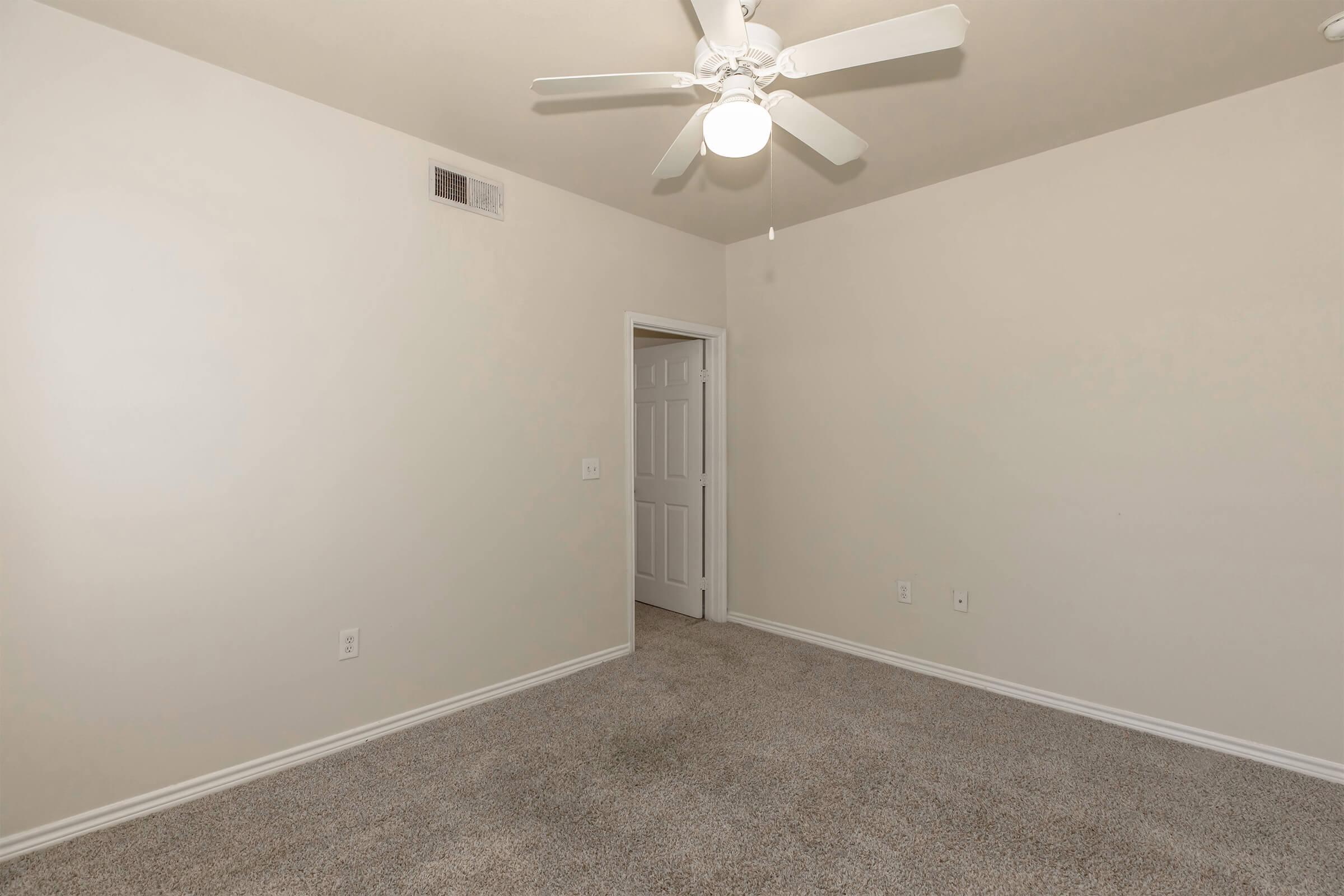 Empty room with light beige walls and a light-colored carpet. A ceiling fan is visible, and there is a door leading to another room on the right. The space is brightly lit and unfurnished, providing a clean and spacious feel.