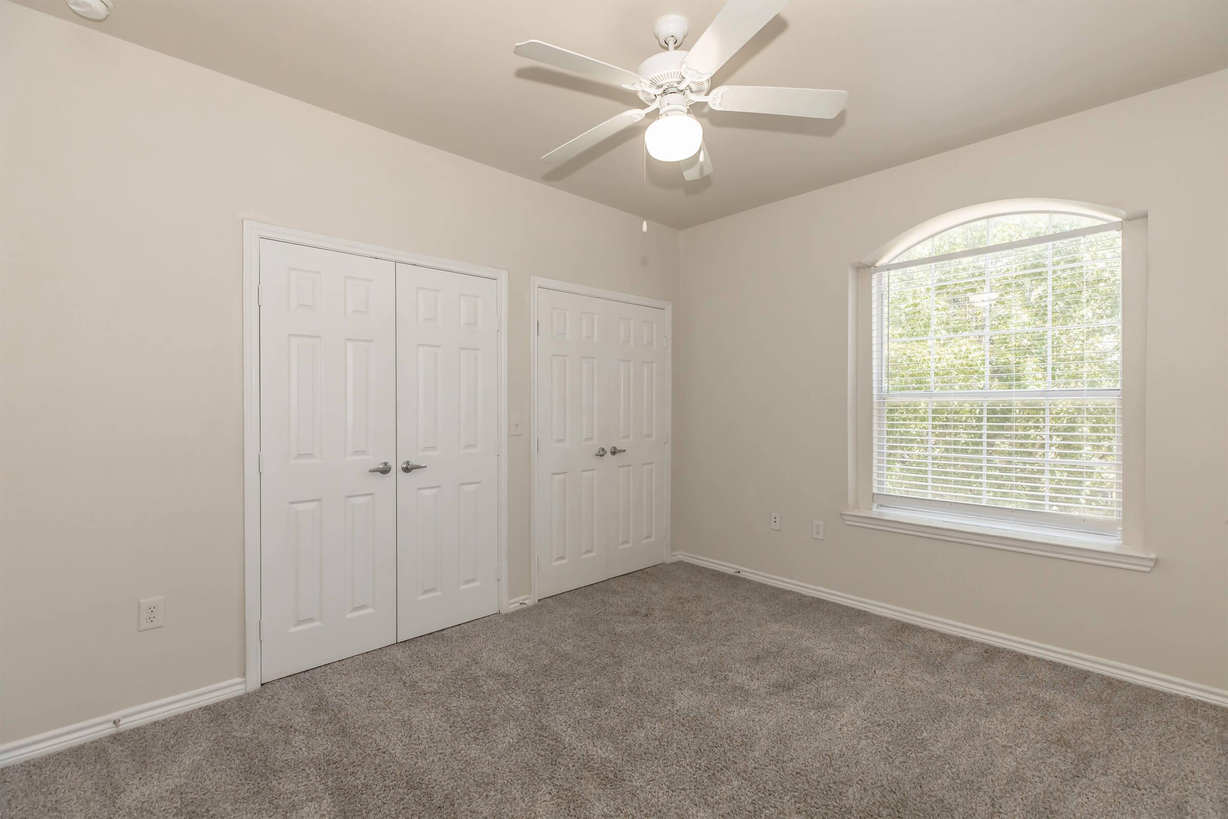A cozy, empty bedroom featuring light beige walls, a ceiling fan with a light fixture, and soft carpet. Two double-door closets are visible on one side, and a window with white blinds allows natural light to brighten the space.