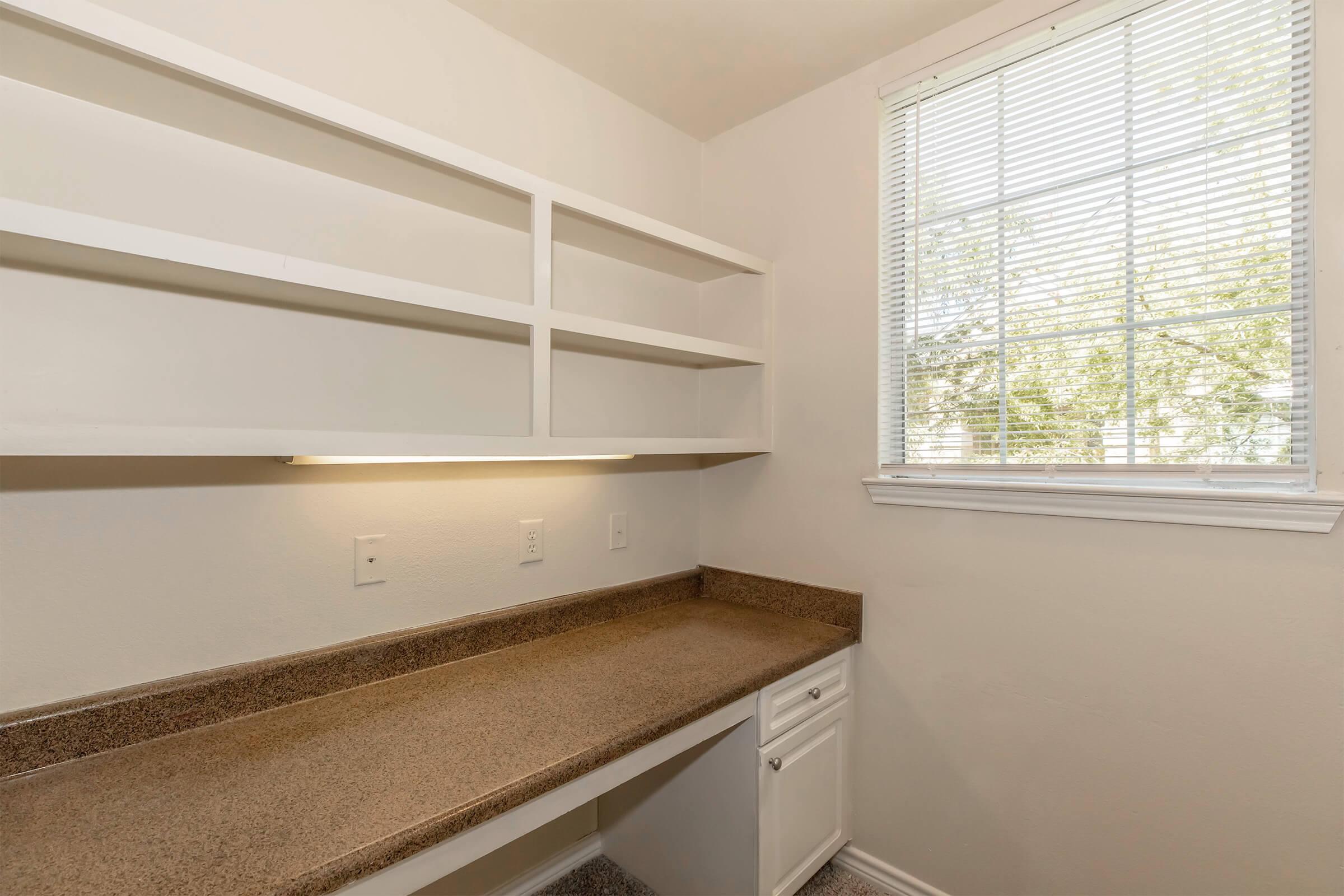 Empty room featuring a long countertop with a built-in cabinet underneath and open shelves above. A window with blinds allows natural light to enter. The walls are painted light and the flooring appears to be carpeted. The space is tidy and uncluttered, suggesting a functional area for work or storage.