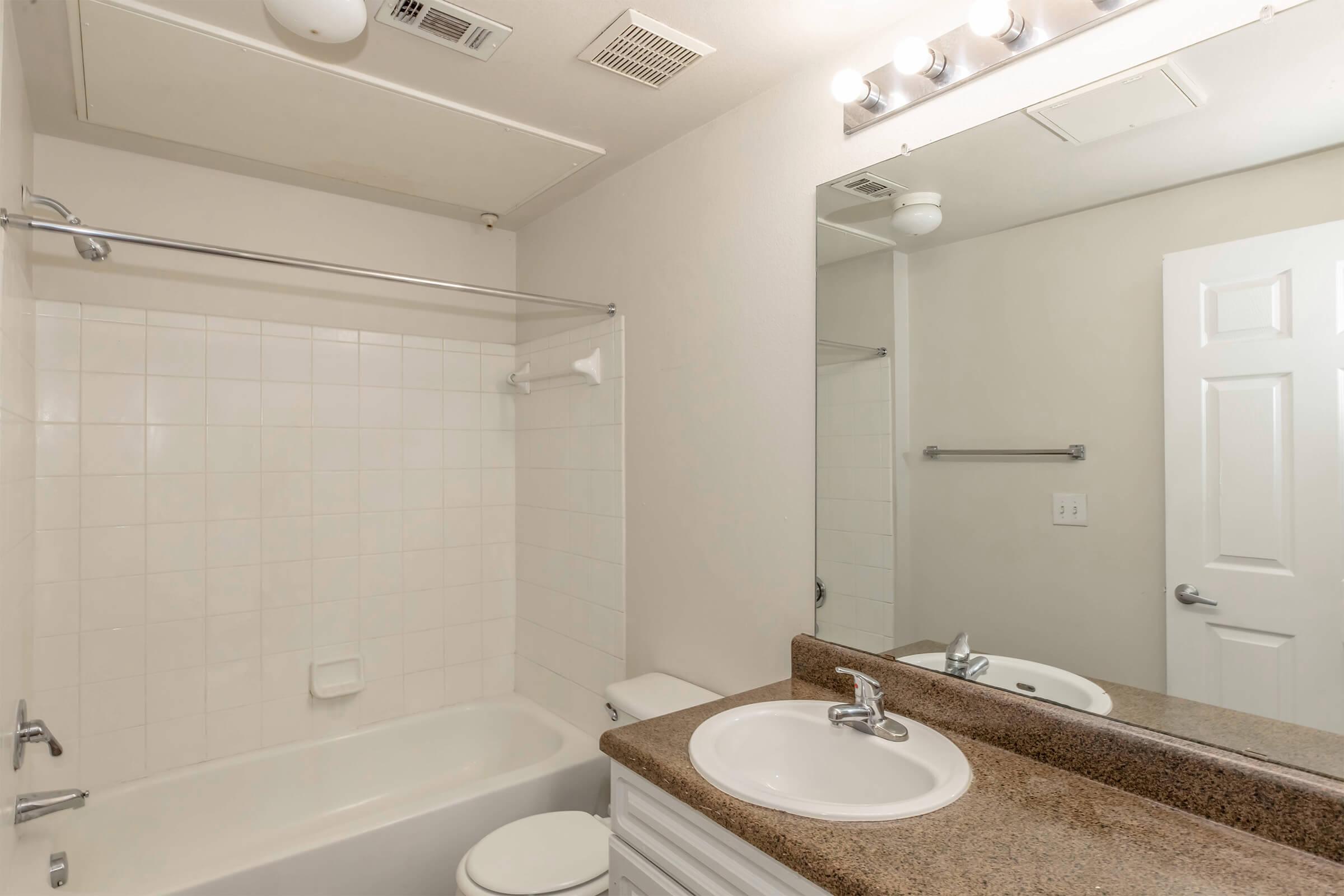 Bright bathroom with a bathtub, shower curtain rod, and a single sink with a granite countertop. A large mirror is above the sink, and there is a light fixture above. The walls are painted a light color, and a door is visible to the right. The bathroom features white tiles and simple fixtures.