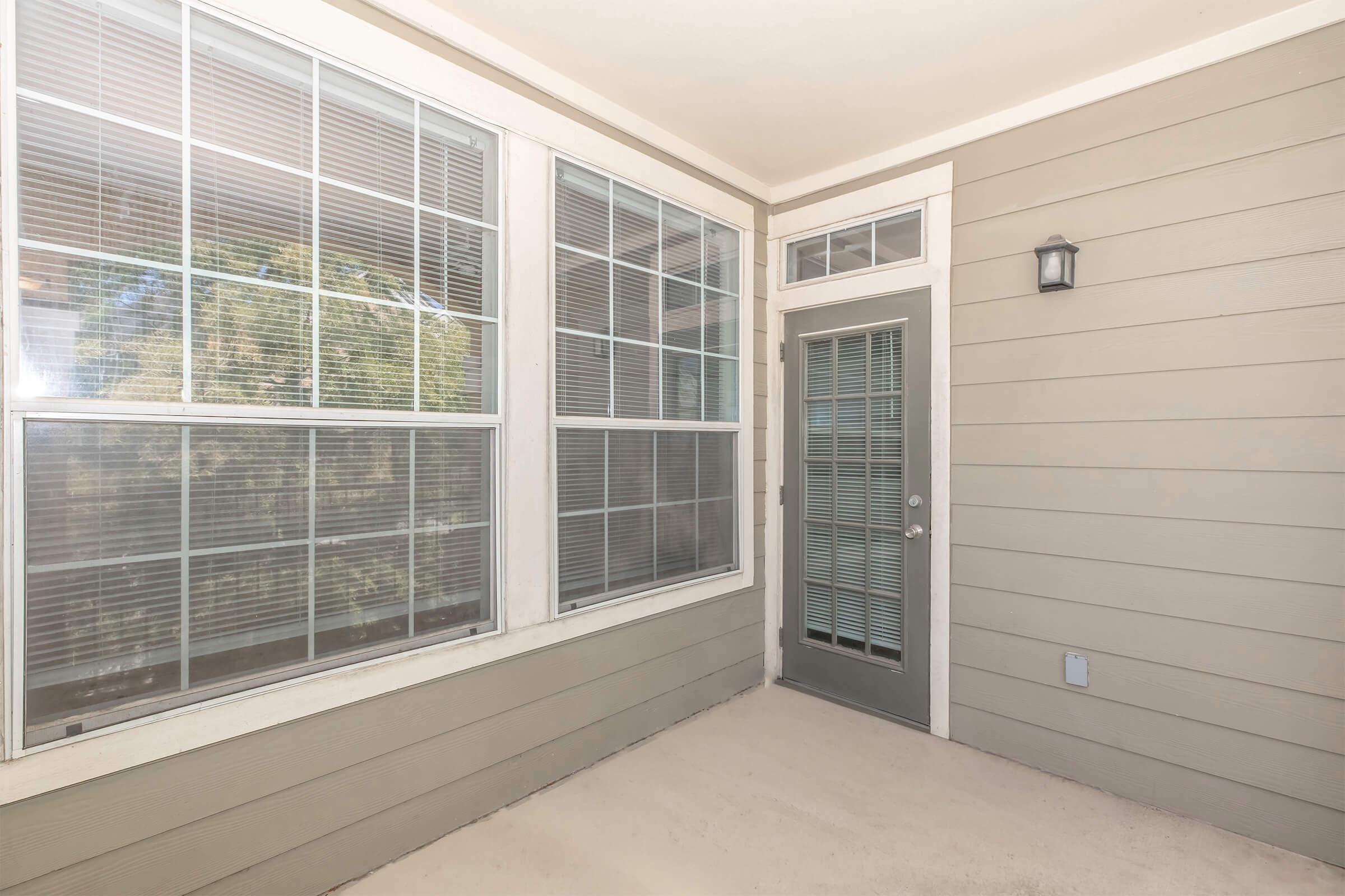 A well-lit and clean porch with large windows featuring multiple panes, a light fixture on the wall, and a glass-paneled door. The walls are painted in light gray, and the floor is plain with a smooth surface, creating an inviting outdoor space. The view outside shows greenery through the windows.