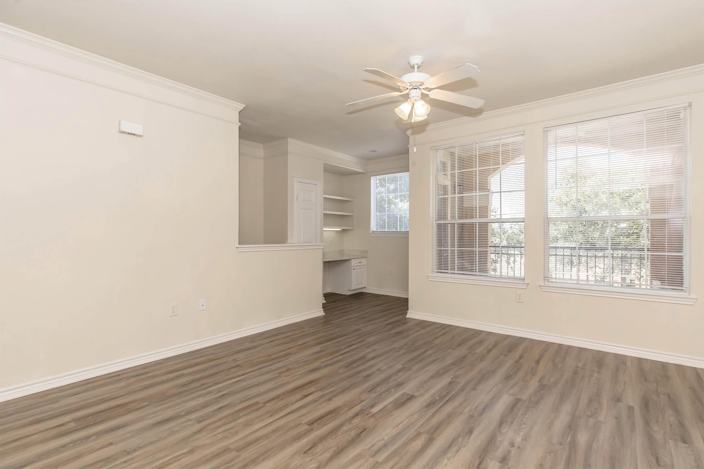 A spacious, empty living room with light-colored walls and a ceiling fan. The floor is covered in light wood laminate. Large windows provide natural light, and there is a small nook with shelves visible in the background. The overall atmosphere is bright and airy.