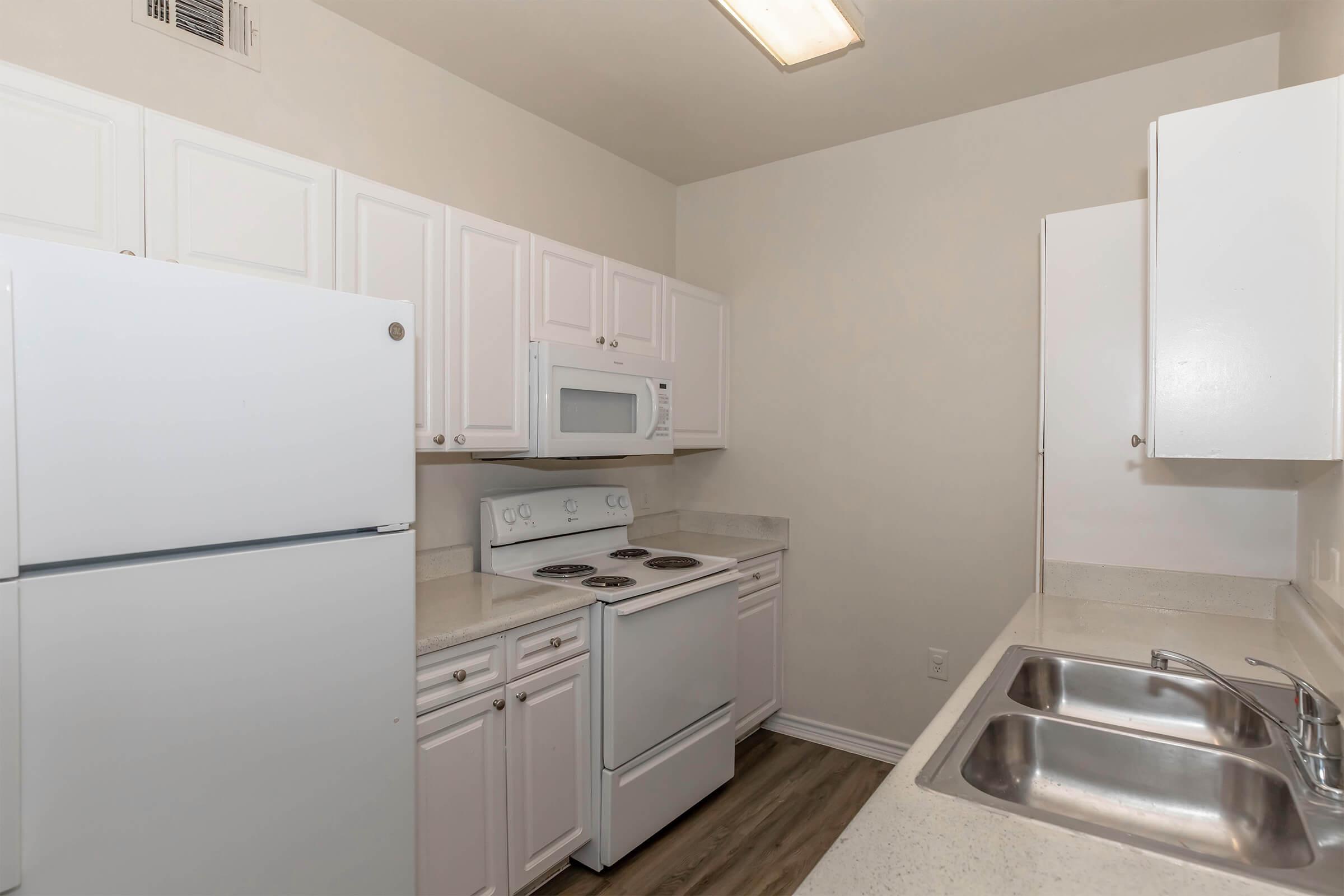 A modern kitchen featuring white cabinets, a white refrigerator, a white microwave, and a white stove. The countertops are light-colored, and there are stainless steel sinks. The flooring is a wood-like laminate, and the walls are painted a neutral color for a clean and bright appearance.