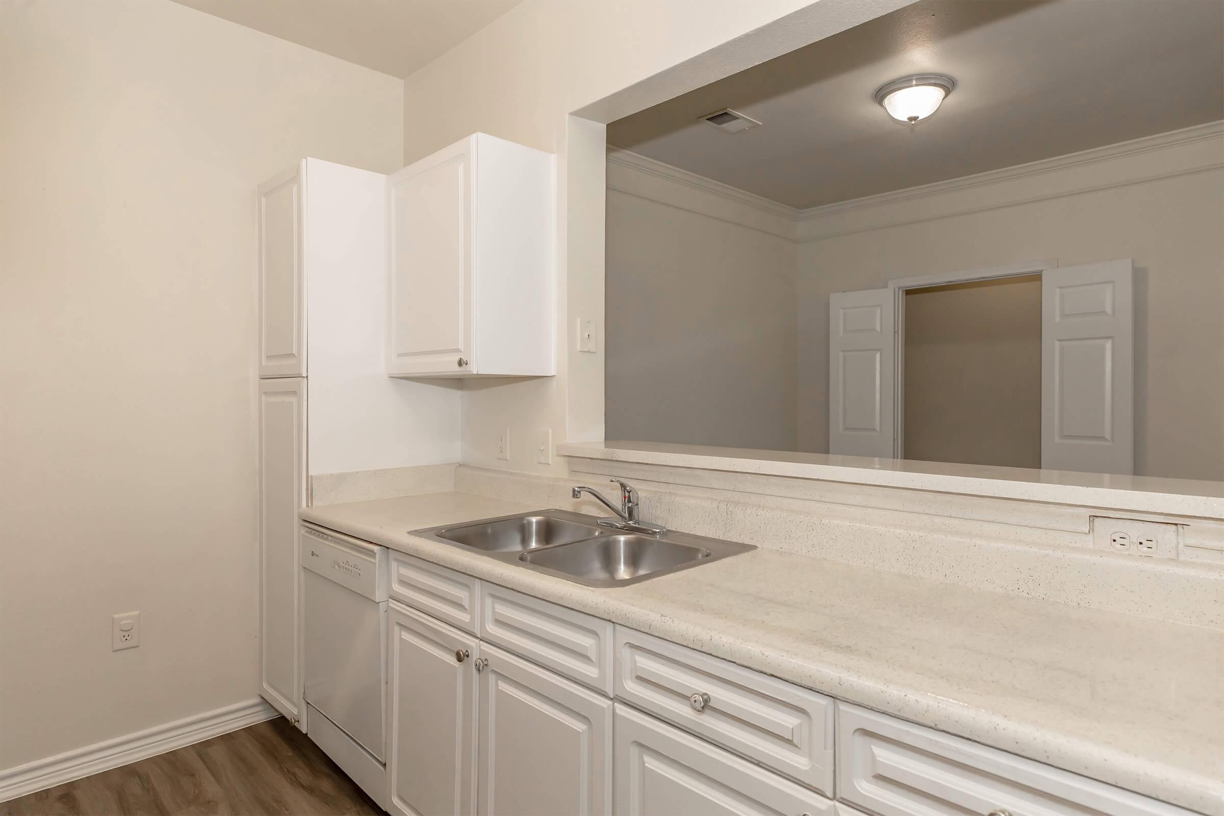 Modern kitchen featuring white cabinets, stainless steel double sink, and a dishwasher. The countertop is light-colored, with a wide opening leading to an adjoining room. Soft, neutral walls and a ceiling light fixture enhance the bright, inviting space. Wood-like flooring completes the contemporary design.