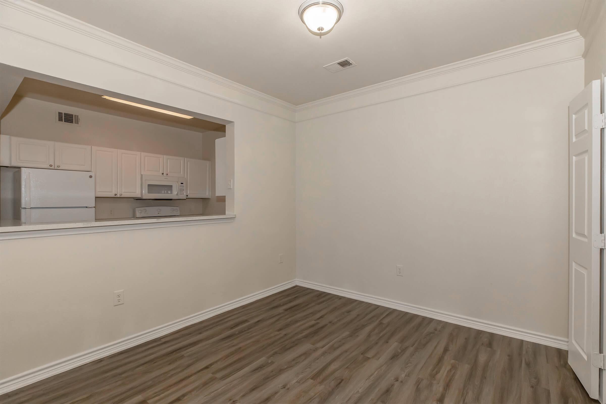 A brightly lit, empty room featuring light-colored walls and a wood-like floor. An open space connects to a kitchen area with white cabinetry, a microwave, and a stove visible through a large archway. The ceiling has a central light fixture, and one wall displays a closed white door.