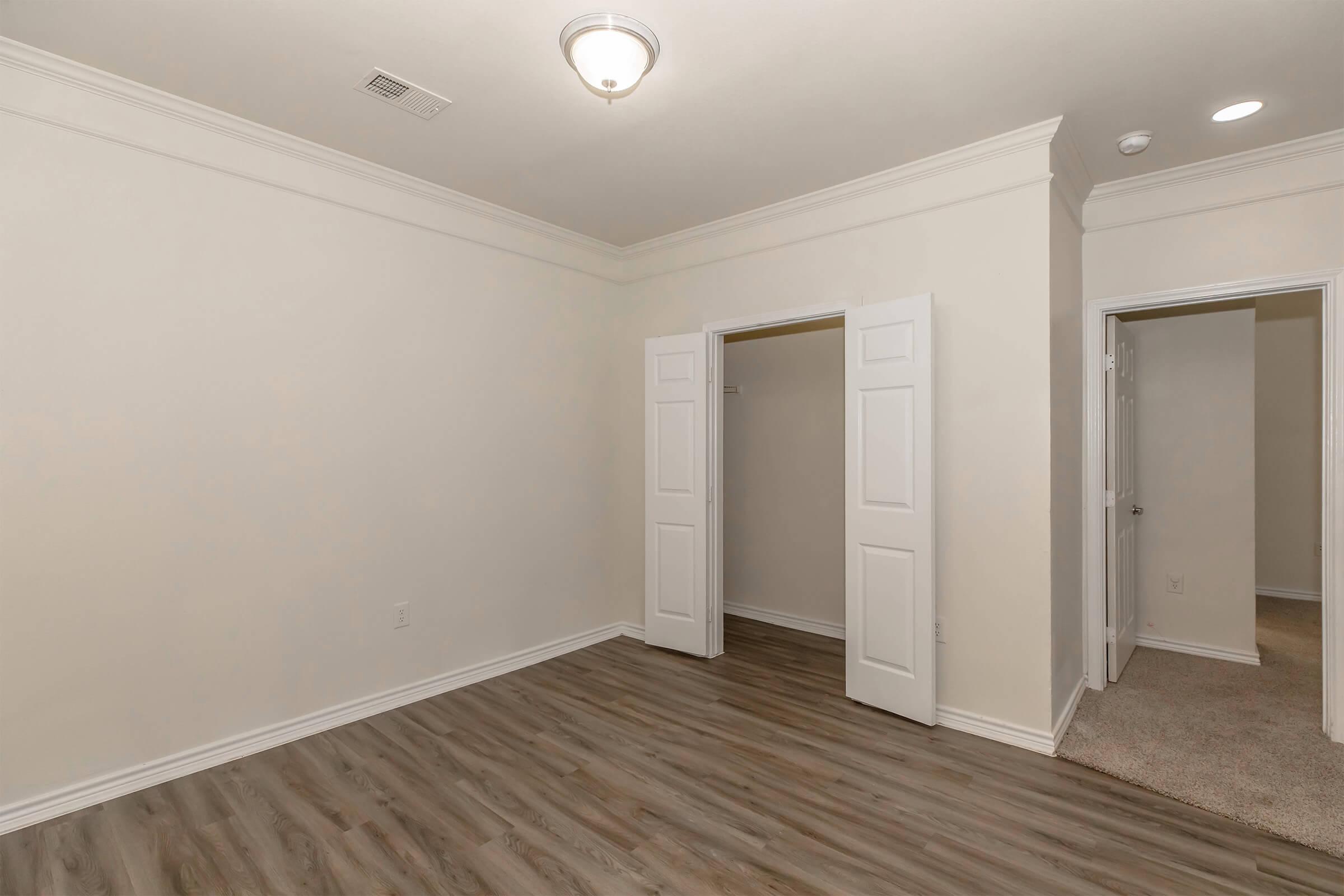 A spacious, empty room with light-colored walls and wooden flooring. It features a ceiling light fixture and two doorways, one leading to a closet and the other to a hallway. The room is well-lit and has a minimalist design, ideal for various decorating options.