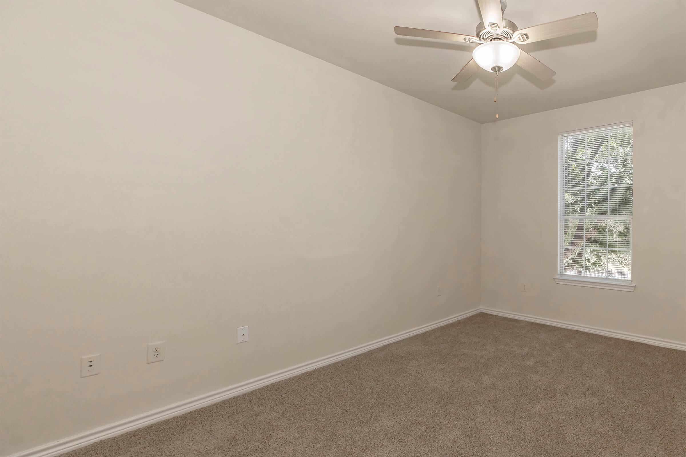 Empty room with light beige walls and carpeted floor. A ceiling fan is installed in the center, and there is a single window with blinds, allowing natural light to filter in. The overall space is unfurnished and appears clean and simplistic.