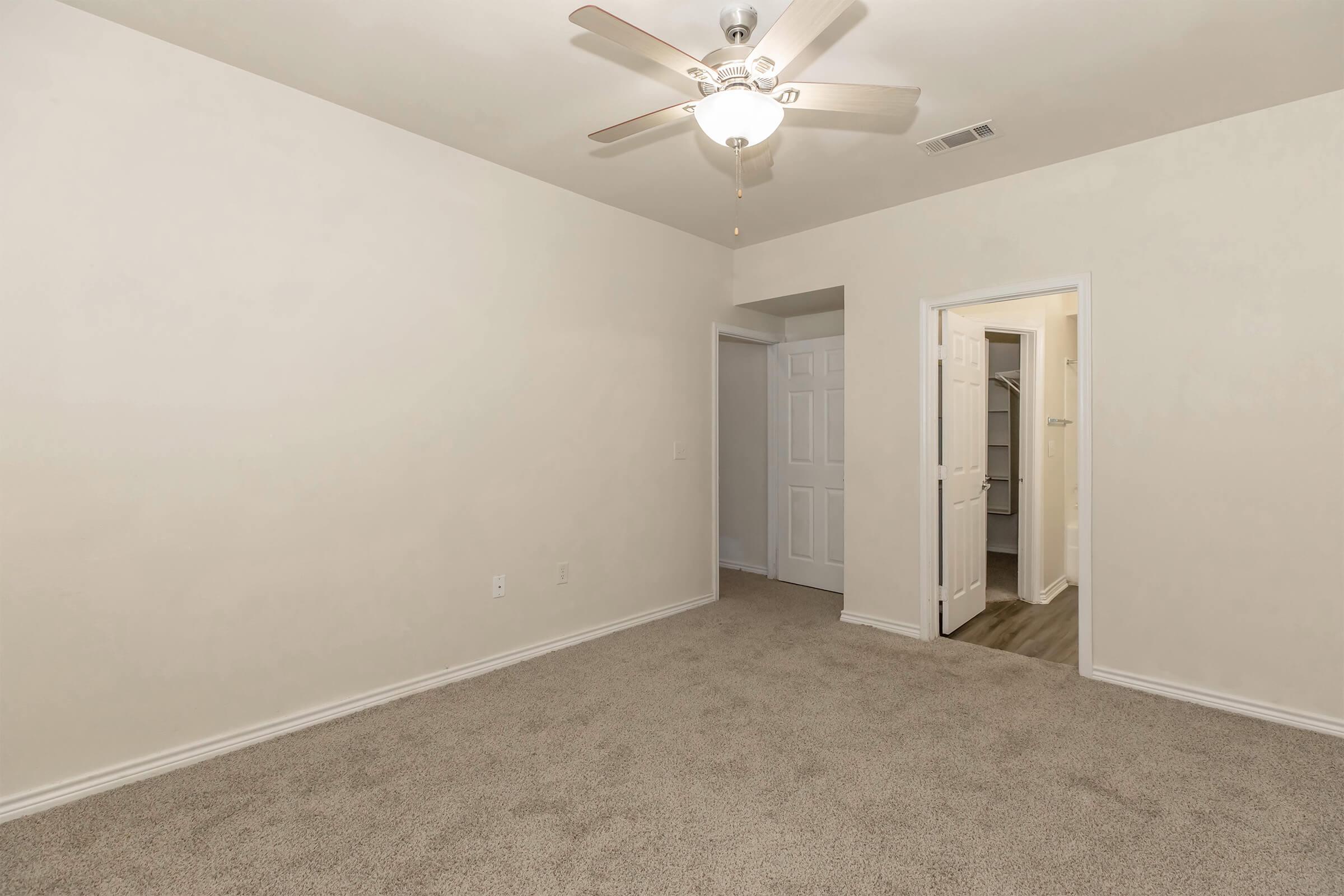 A spacious, carpeted room with light beige walls and a ceiling fan. It features an open door leading to a closet area and another door that leads to a hallway. The floor is a light wood laminate, enhancing the overall brightness of the space. Ideal for use as a bedroom or office.