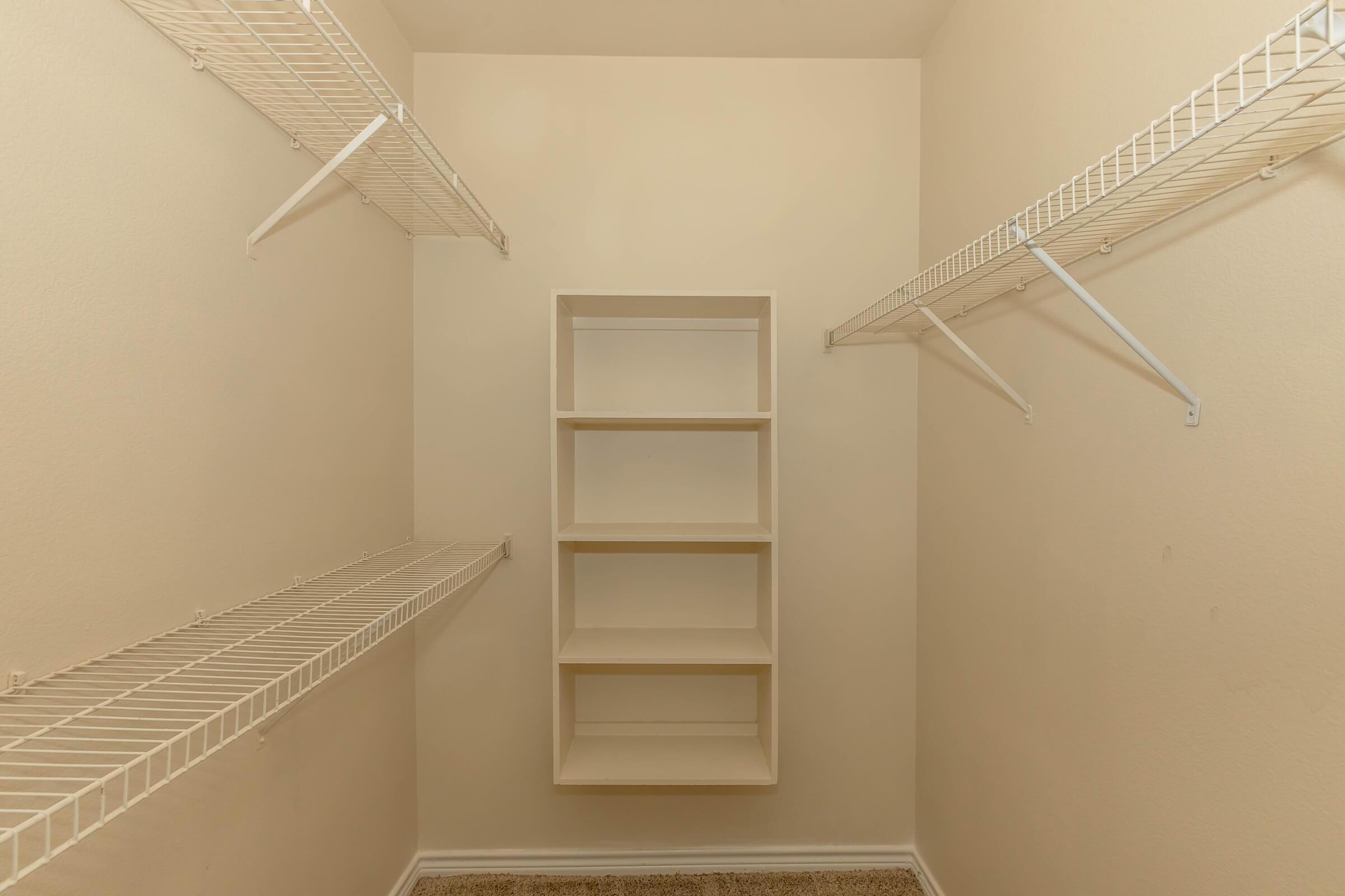An empty walk-in closet featuring beige walls and a carpeted floor. The space includes three wire shelves on either side and a central open shelving unit with four compartments, creating an organized storage area. The overall appearance is minimalist and well-lit.