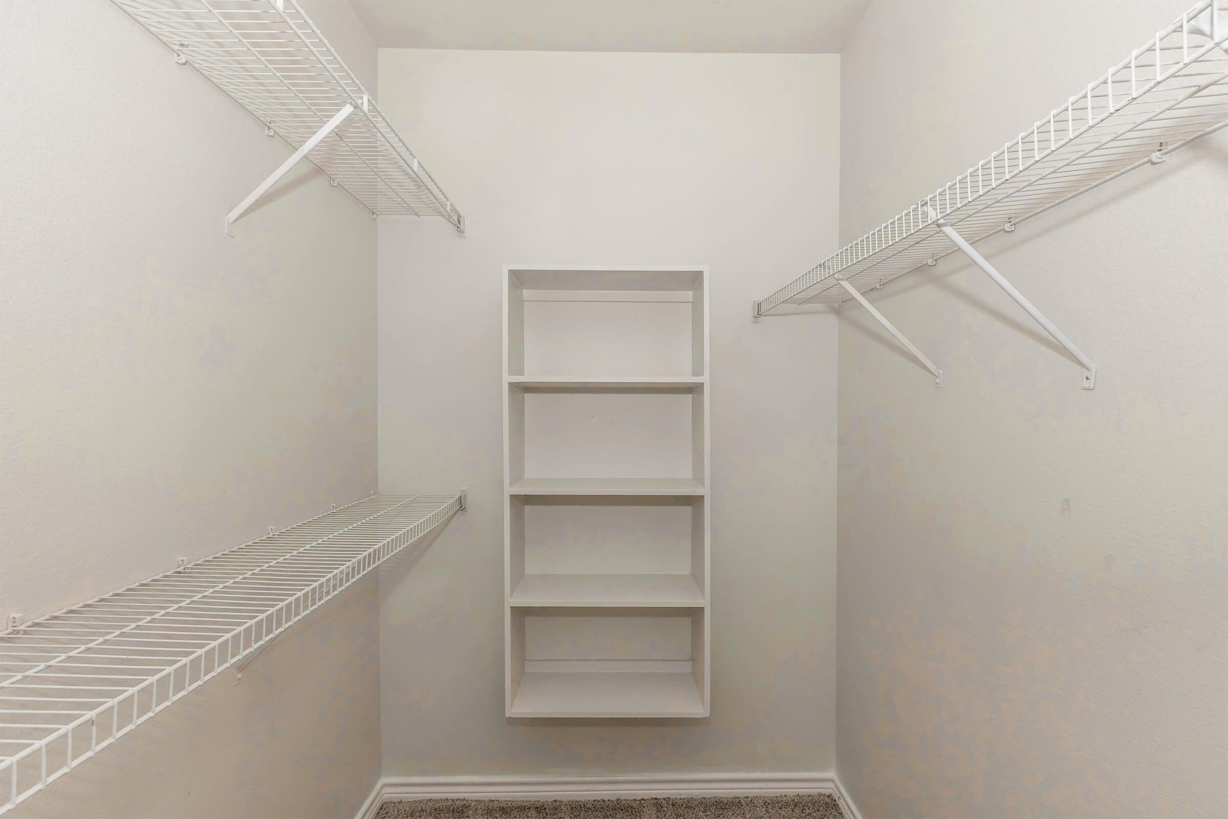 A small empty closet with white wire shelves on both sides and a central shelving unit. The walls are painted in a light color, and the floor is covered in neutral carpet. The space is tidy and offers potential for organization and storage.