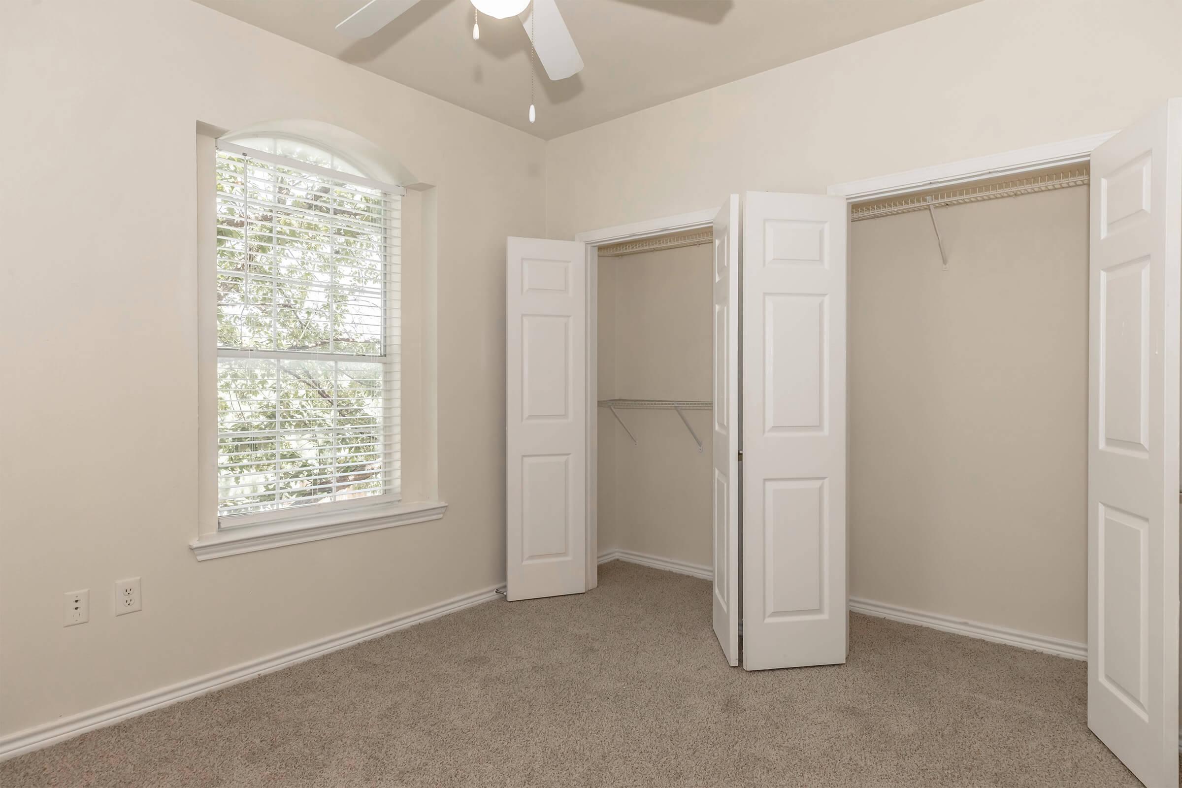 Empty room with beige walls and light carpeting, featuring a window with blinds letting in natural light. There are two double doors leading to an empty closet on one side, and a ceiling fan is visible.