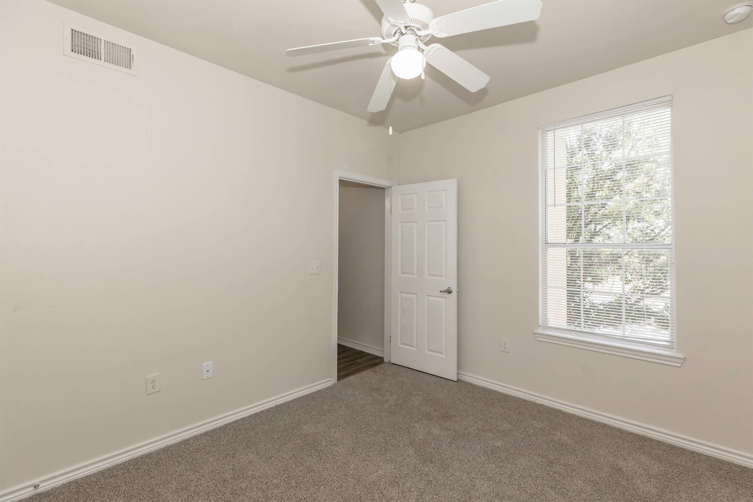 A small, empty bedroom with light beige walls and carpet. The room features a ceiling fan, a window with blinds allowing natural light, and a door leading to another room. The space appears clean and well-maintained, suitable for personal decoration or furniture placement.