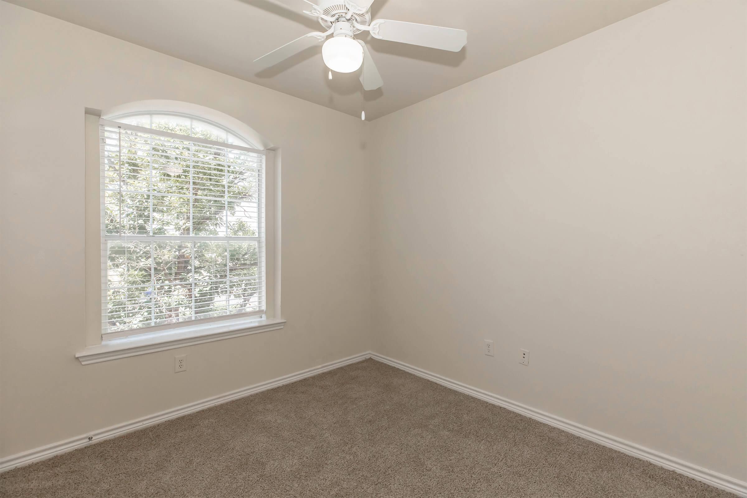 A vacant room with light beige walls and a ceiling fan. There is a window with blinds allowing natural light to enter, and the floor is covered with light-colored carpet. The room appears clean and ready for furniture.