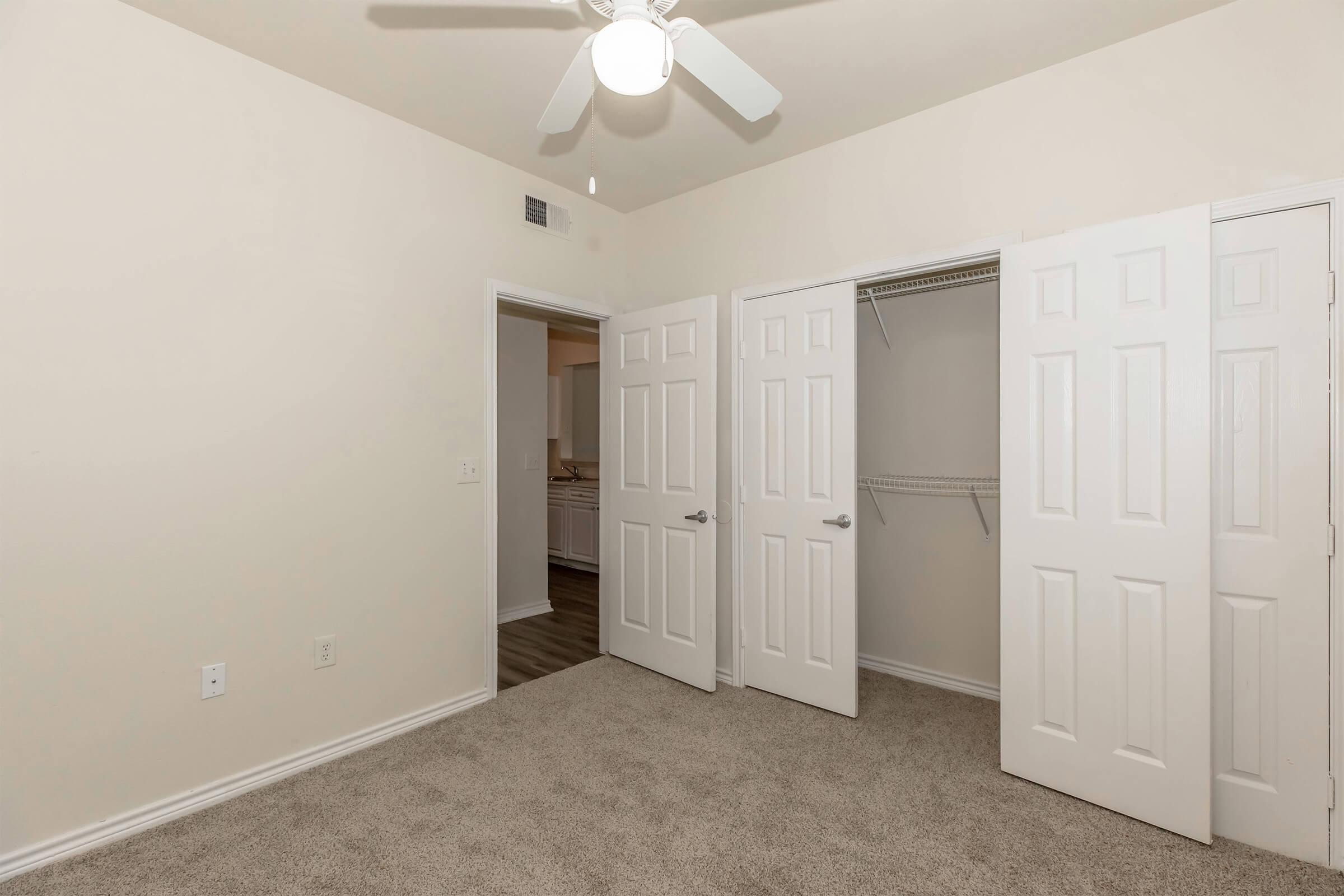 A well-lit bedroom with beige walls and carpet. The room features a ceiling fan and two closet doors that are slightly open, revealing a closet space. A doorway leads to another room in the background, indicating a spacious layout. The overall ambiance is clean and inviting.