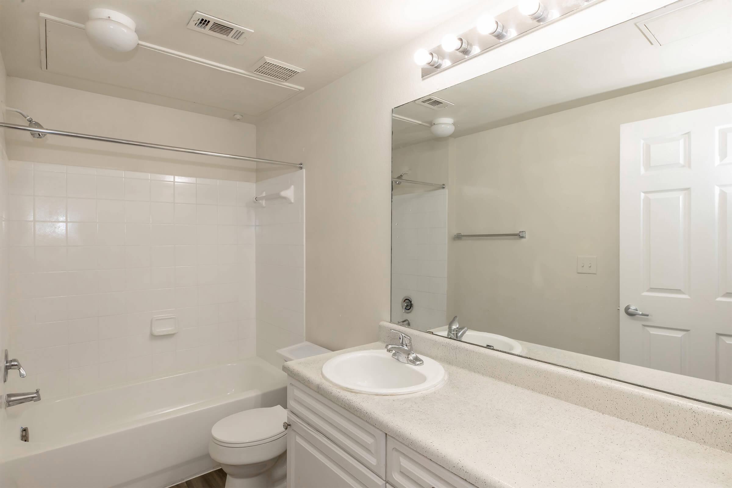 A clean, neutral-colored bathroom featuring a white bathtub, toilet, and sink with a light-colored countertop. The mirror above the sink is well-lit by vanity lights, and the walls are painted in a soft hue. The room has a simple, uncluttered design with a shower curtain rod but no curtain.