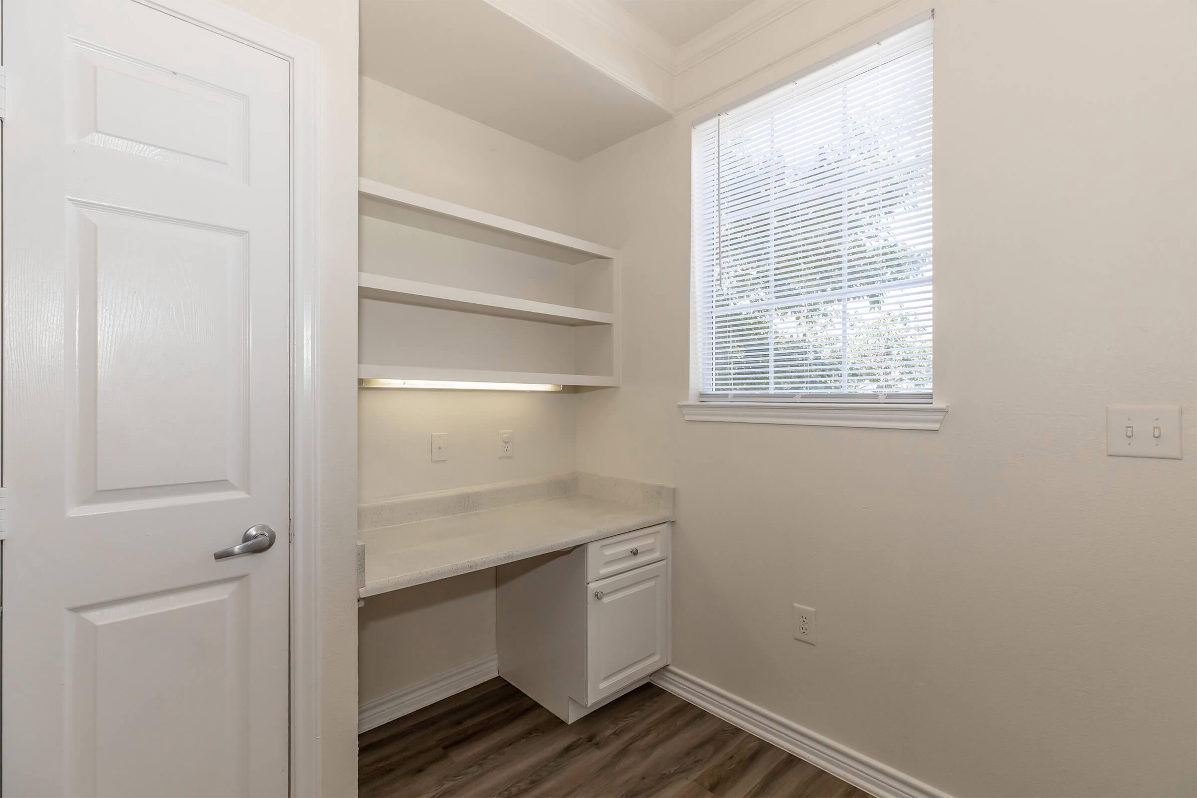 A small, well-lit room featuring a white desk with a built-in drawer, open shelves above, and a window with blinds allowing natural light to enter. The walls are painted white, and the flooring is a light-colored wood laminate. The space is clean and organized, suitable for a home office or workspace.