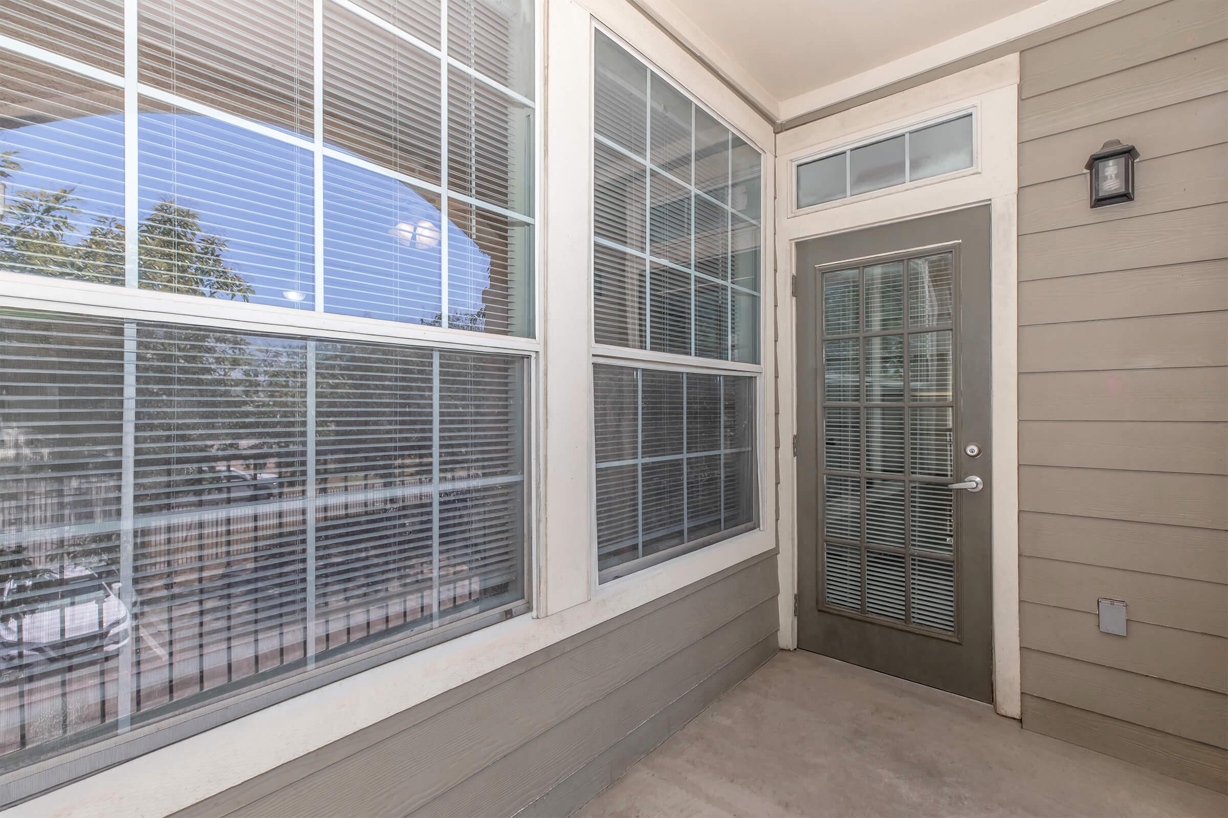 A covered outdoor patio area with large windows featuring white blinds. The main door is gray with a glass panel and a small light fixture above. The walls are beige with light trim. Trees are visible outside through the windows, indicating a peaceful environment.