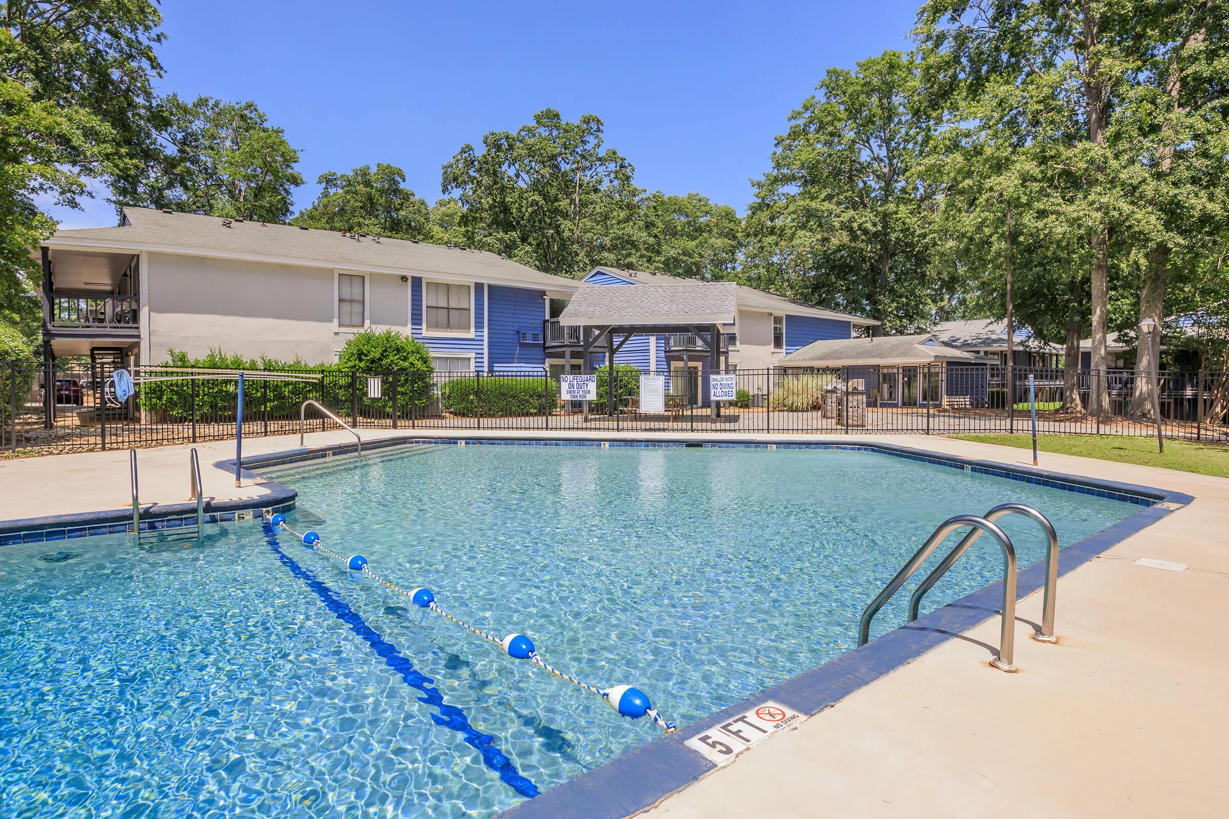 A clear swimming pool with a shallow end marked by a sign. Surrounding the pool are trees and residential buildings, all set under a bright blue sky. The area is well-maintained with lounge chairs and fencing around the pool.