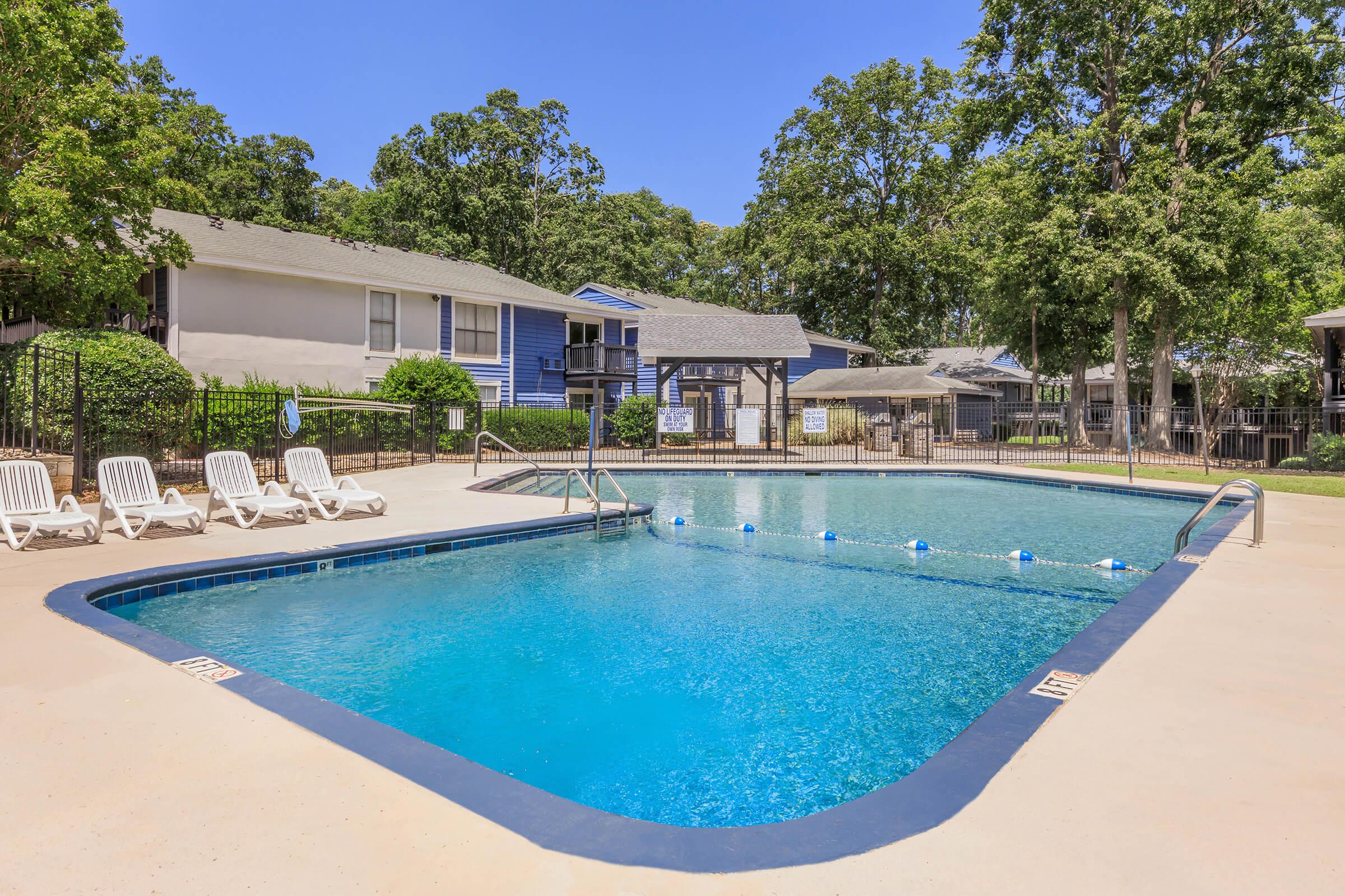A sparkling blue swimming pool surrounded by a concrete deck, with white lounge chairs lined up on one side. In the background, there are two-story apartment buildings under a clear blue sky and trees providing shade. A few pool floats are visible in the water.
