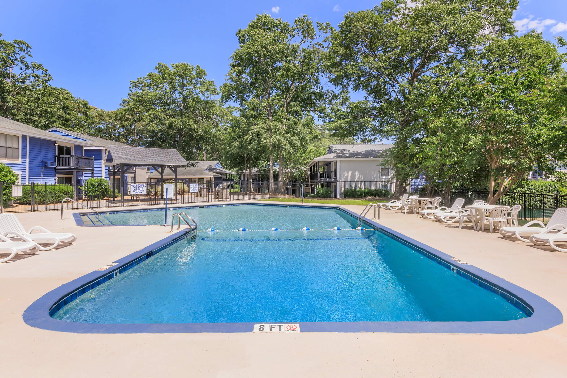 A bright and inviting outdoor pool area surrounded by trees. The rectangular pool is clear and calm, with a depth marker indicating 8 feet. Several white lounge chairs are positioned around the poolside, and cozy buildings are visible in the background beneath a clear blue sky.