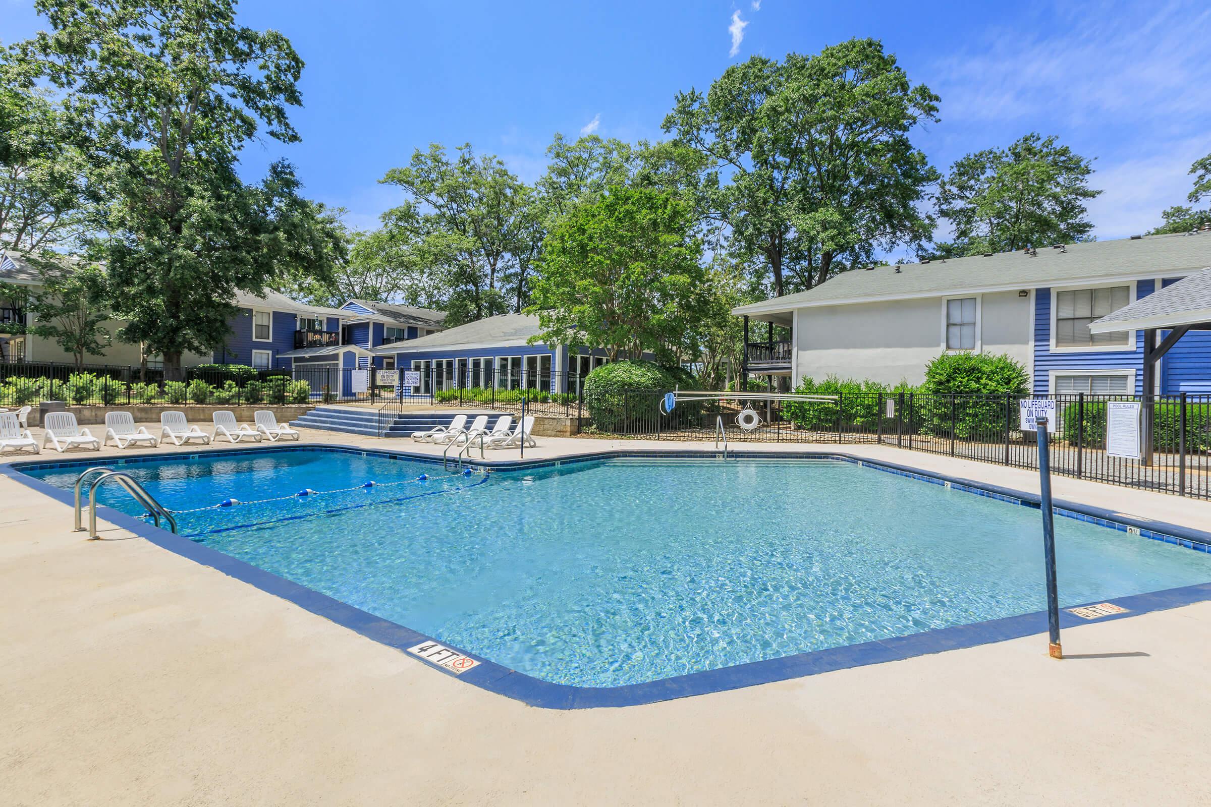 A tranquil outdoor swimming pool surrounded by lounge chairs, trees, and apartment buildings under a clear blue sky. The pool features a clean, inviting surface with clear water, and there are safety signs visible nearby. The area is well-maintained and offers a relaxing atmosphere for residents.