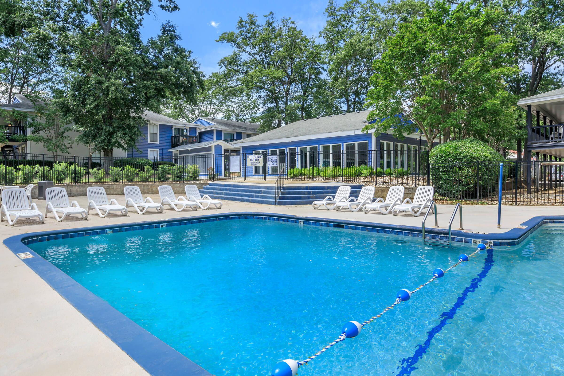 A clear blue swimming pool surrounded by white lounge chairs, with lush green trees and landscaped areas in the background. A building with blue siding is visible behind the pool, and the area is sunny with a blue sky above.