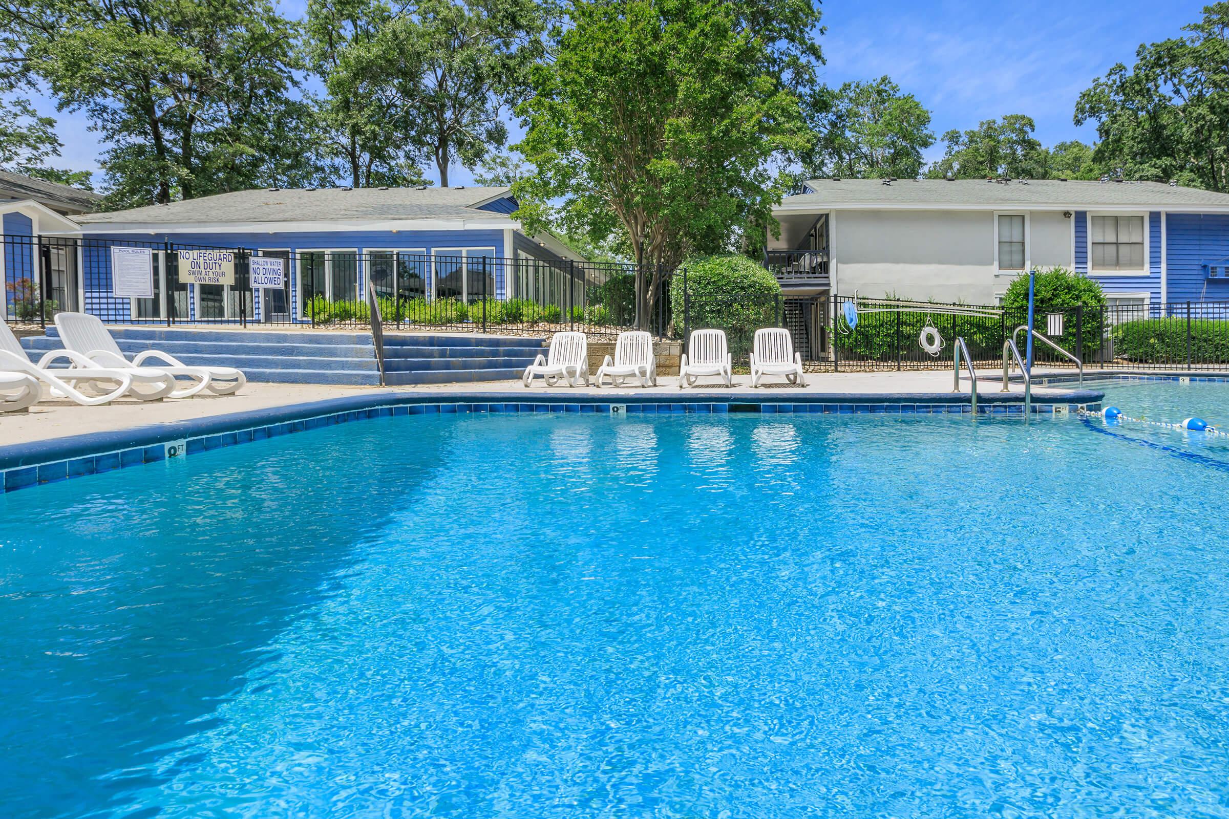 A sparkling blue swimming pool surrounded by lounge chairs. In the background, there are two buildings with green landscaping and trees. The area is well-maintained, offering a relaxing atmosphere for outdoor enjoyment. Bright blue sky adds to the inviting scene.