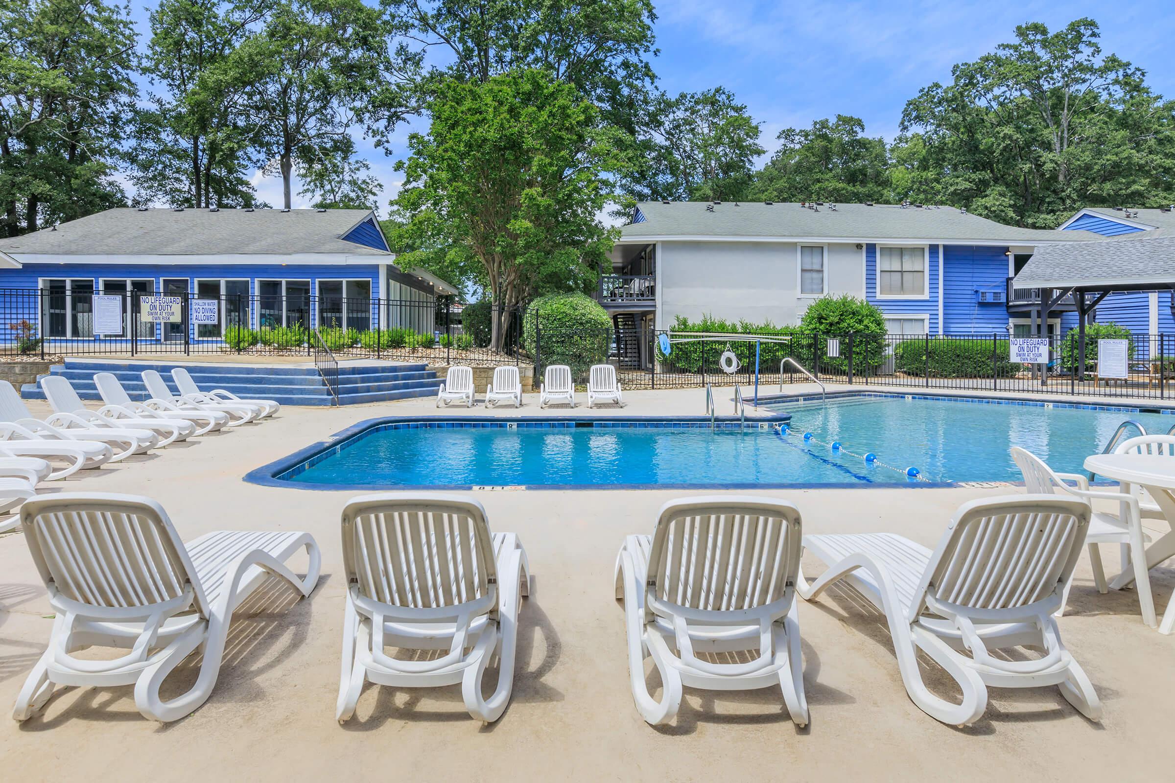 A swimming pool surrounded by white lounge chairs, with green trees and blue buildings in the background. The pool area is well-maintained, featuring a clear blue water surface and a calm, inviting atmosphere. Ideal for relaxation and leisure.
