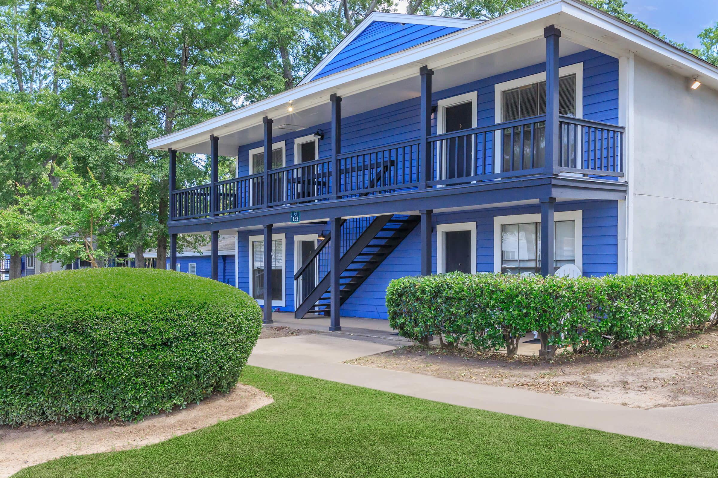 Two-story, blue apartment building featuring a balcony and staircase. Surrounding greenery includes a manicured hedge and grassy area, with a pathway leading to the entrance. The building has large windows and a welcoming exterior.
