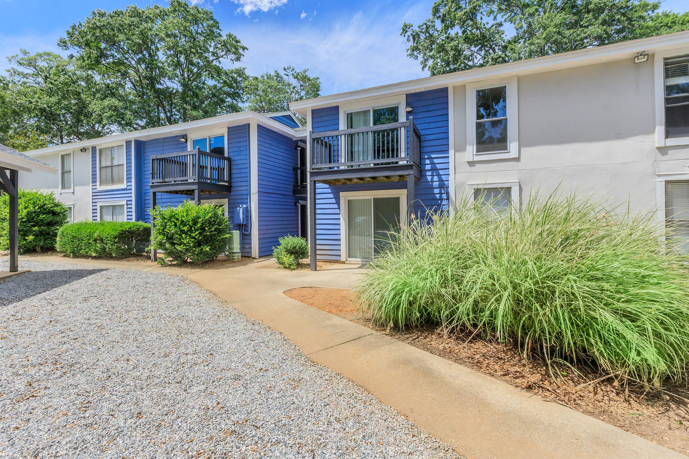 A picturesque view of a multi-unit residential building featuring blue and white exterior colors. The scene includes pathways lined with gravel, greenery, and a clear blue sky with scattered clouds, creating a welcoming atmosphere. Balconies on the upper level add to the charm of the property.