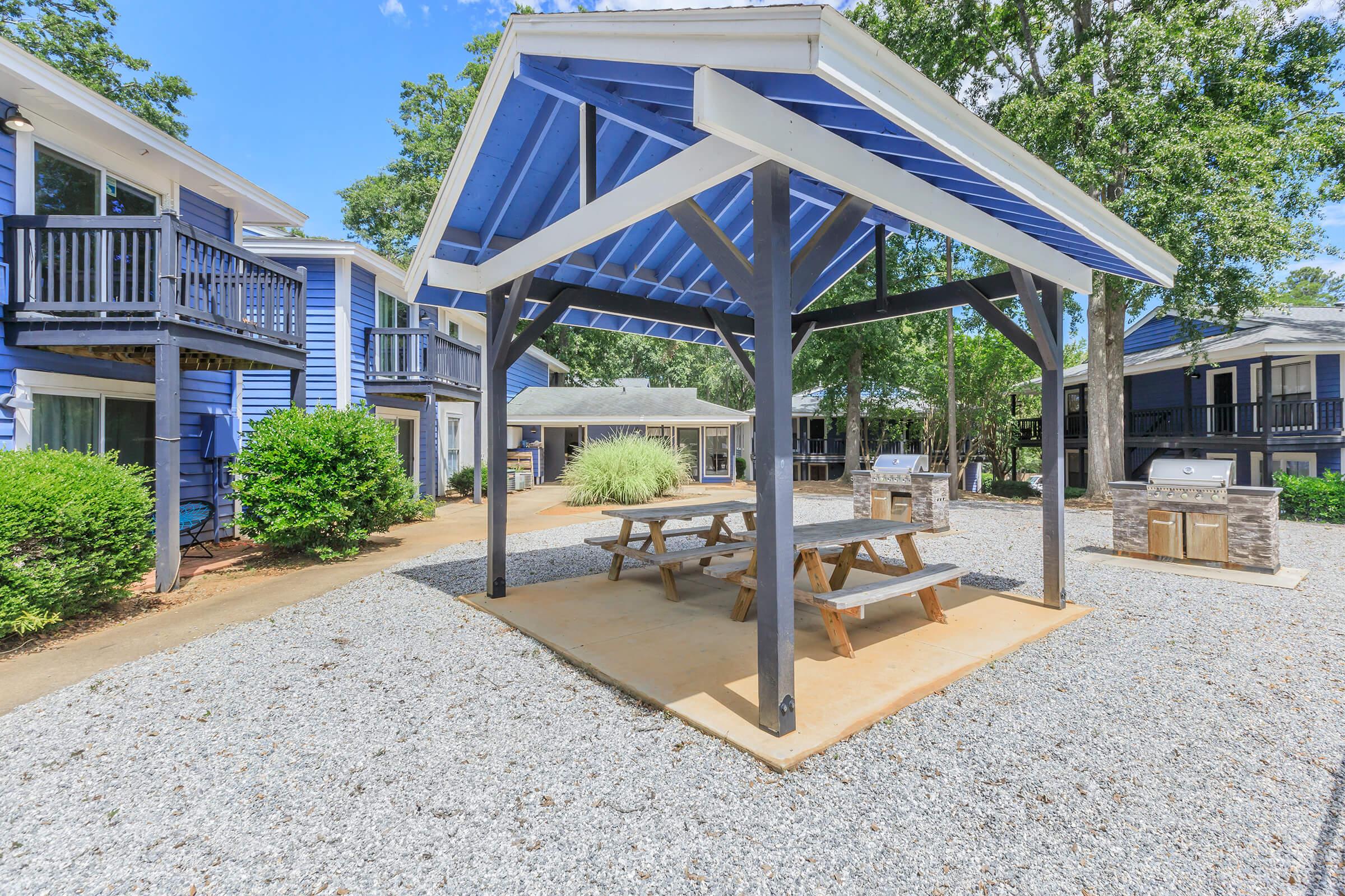 A picnic area featuring a large gazebo with a blue roof, surrounded by gravel and green shrubs. There are picnic tables underneath the gazebo, and nearby, additional seating and grilling stations are visible. The scene is bright and sunny, showcasing a well-maintained outdoor space.