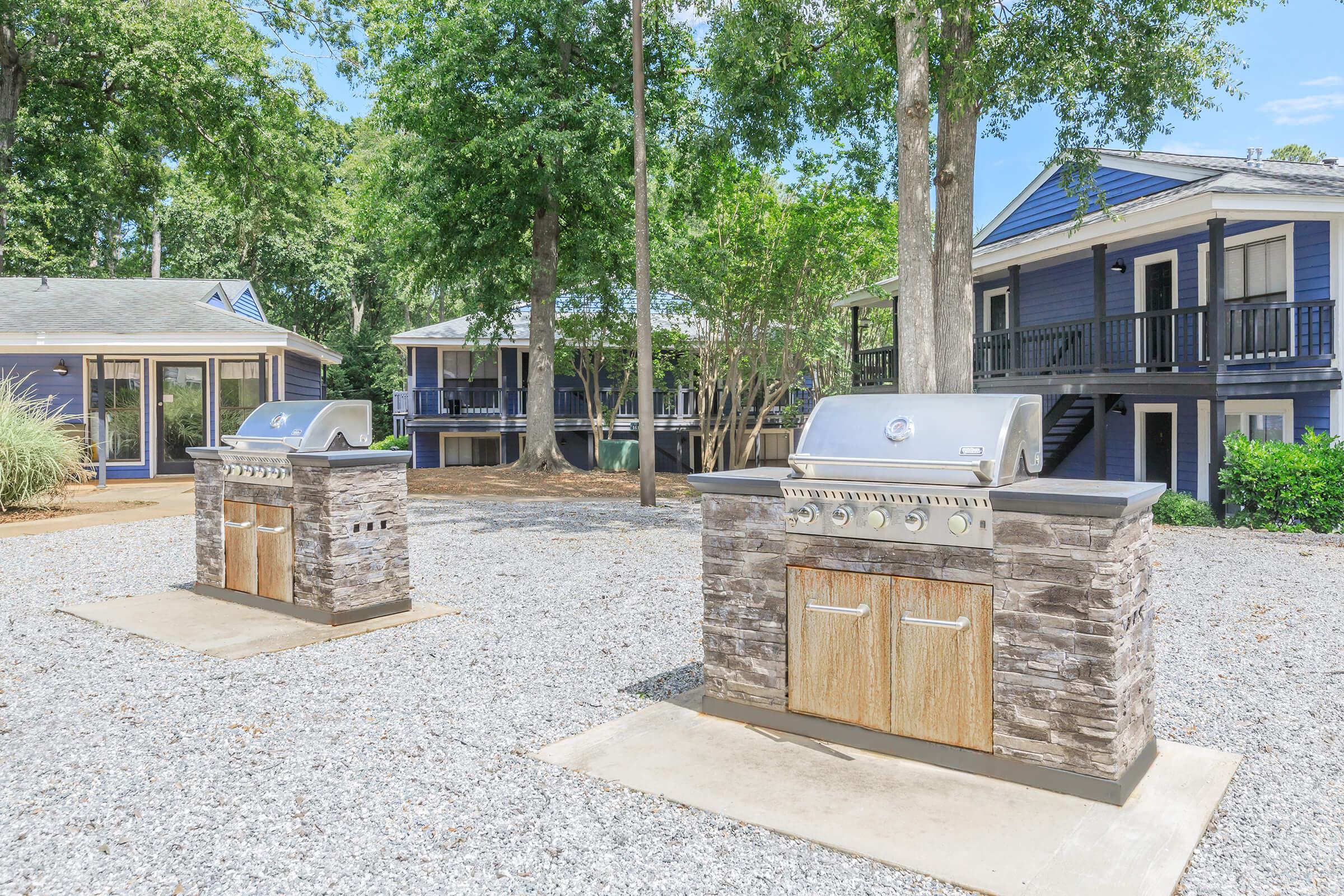 Two outdoor grills built into stone structures are positioned on a gravel area. Surrounding greenery includes trees, and there are blue and white buildings in the background, indicating a communal or recreational space.