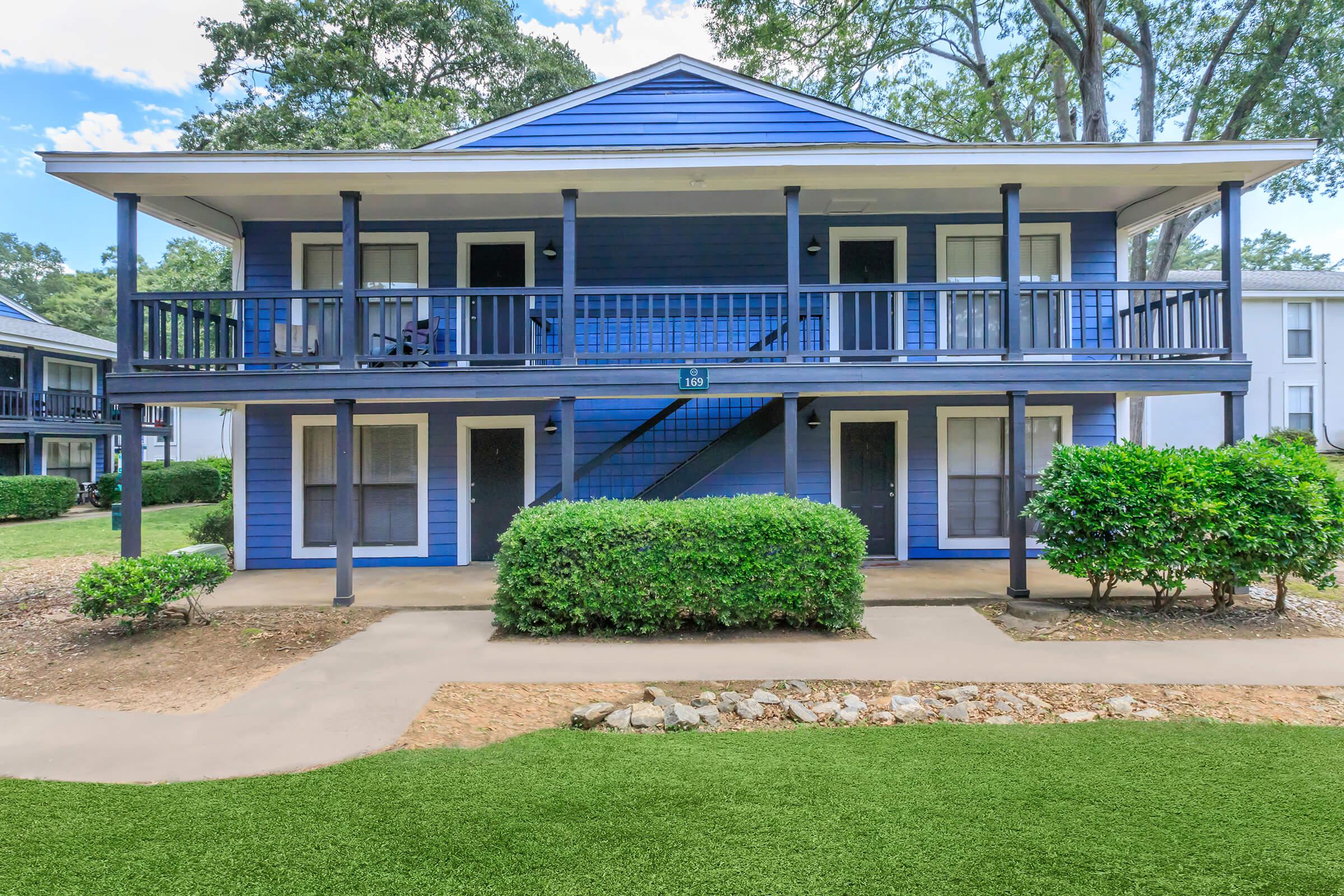 A two-story blue apartment building with a staircase in the center. Each level features two balconies with black railings and multiple doors. The surrounding area has green shrubs and well-maintained grass. Bright skies with scattered clouds are visible above.