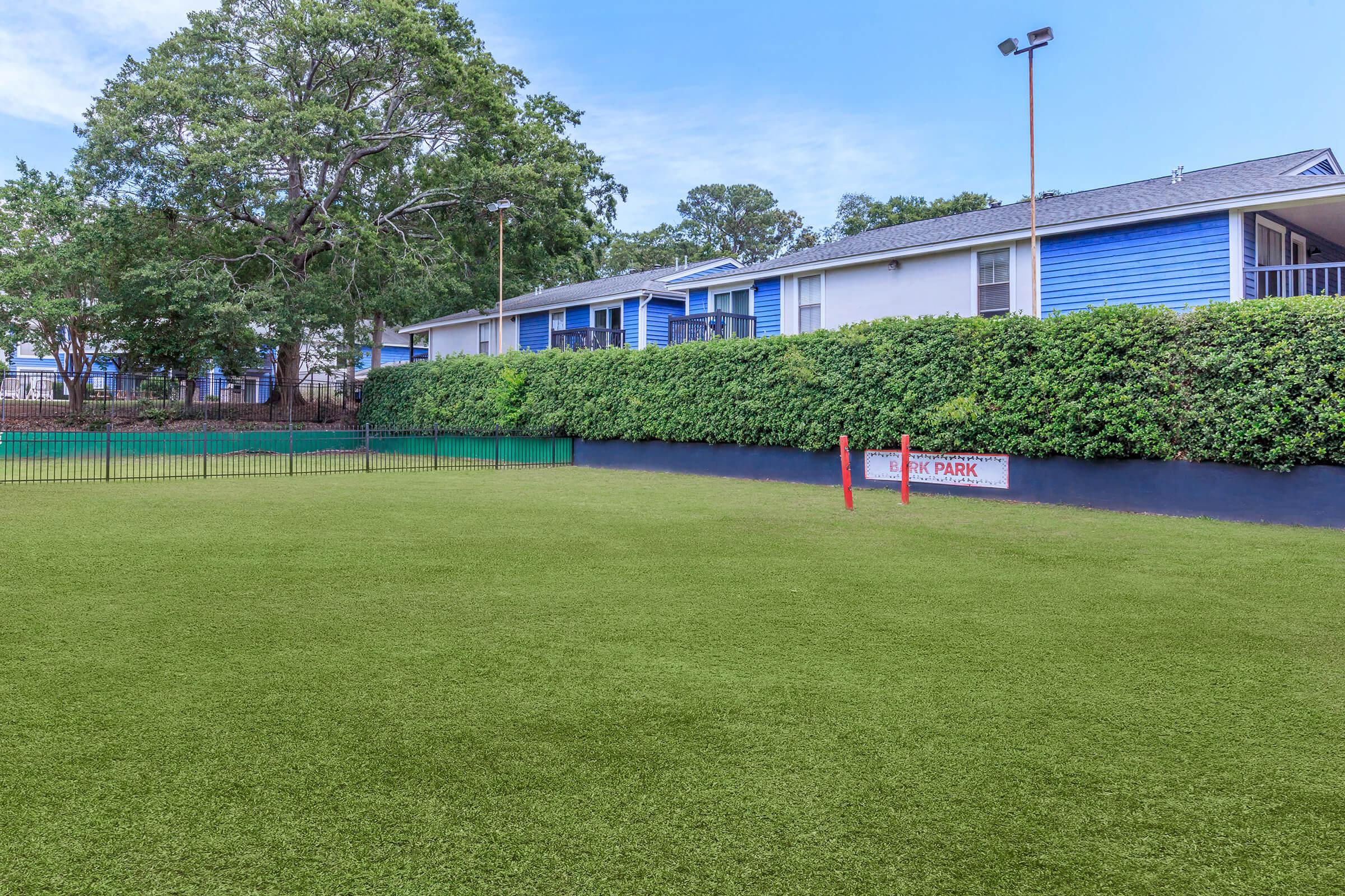 A well-maintained dog park featuring artificial turf, surrounded by green hedges and a fence. In the background, there are residential buildings with blue exteriors. A sign reading "DOG PARK" is visible near the entrance, indicating the designated area for pets. The scene is bright and inviting.