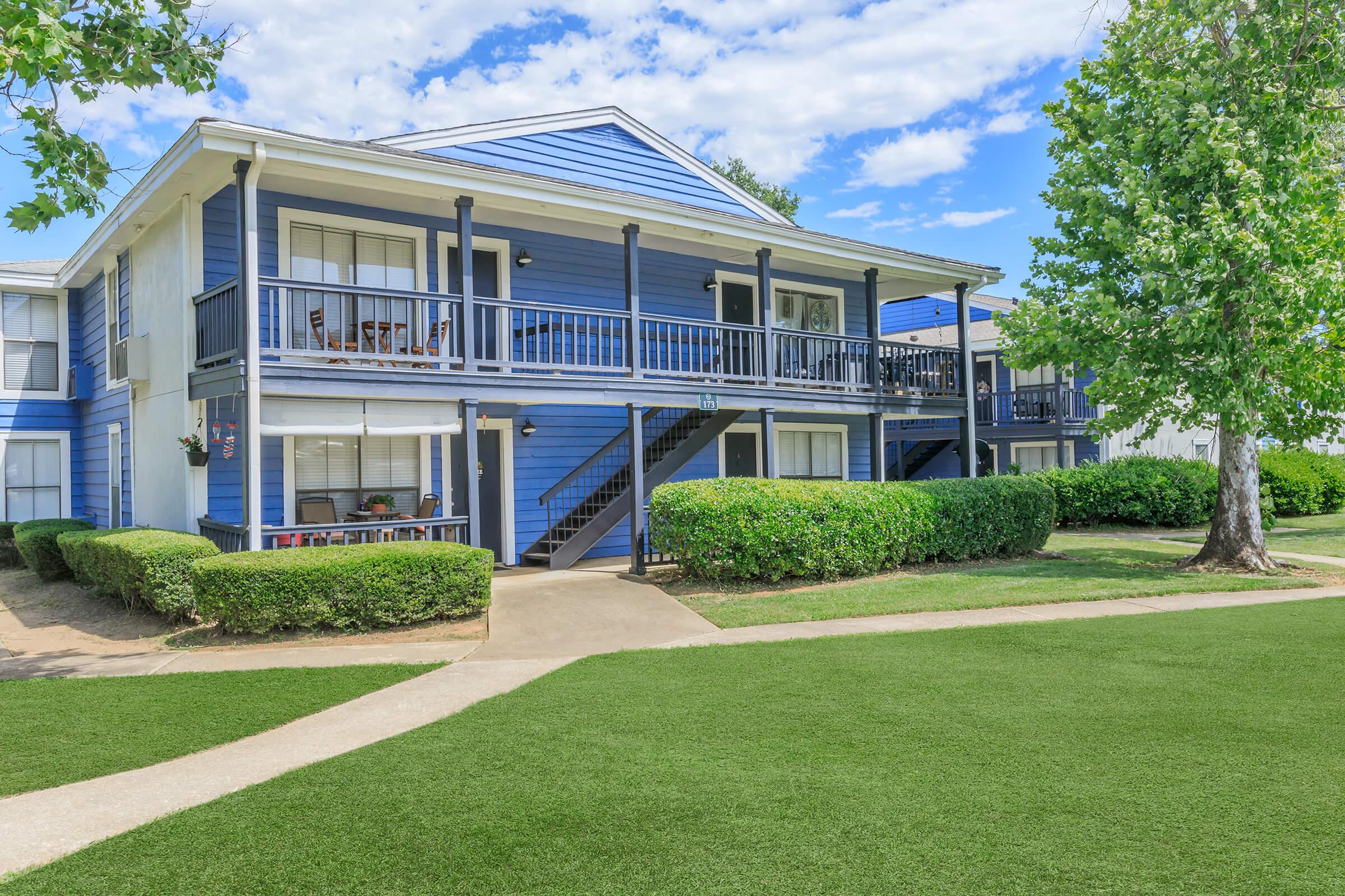 Two-story blue apartment building with balconies and a well-maintained lawn. Surrounding greenery includes neatly trimmed bushes and a tree. A pathway leads to the entrance, and the sky is bright with scattered clouds.