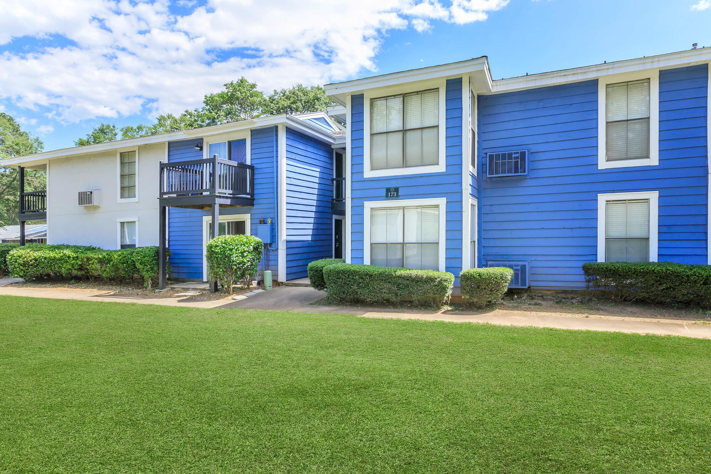 Two-story blue apartment building with white trim, featuring several windows and a balcony. The building is surrounded by well-maintained greenery and grass under a bright blue sky with scattered clouds.