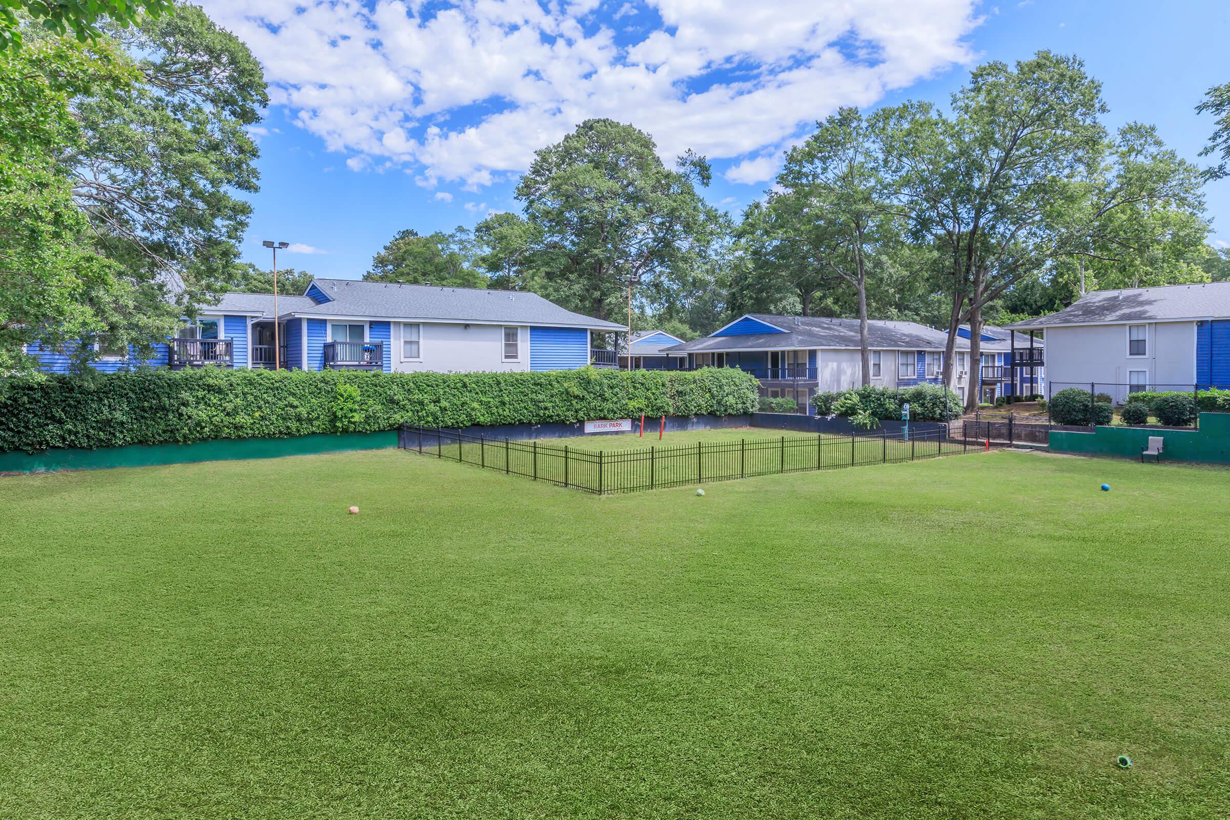 A sunny view of a well-maintained green lawn surrounded by several residential buildings. The lawn is enclosed with a black fence, and there are small trees and hedges nearby. The sky is blue with scattered clouds, creating a pleasant atmosphere.