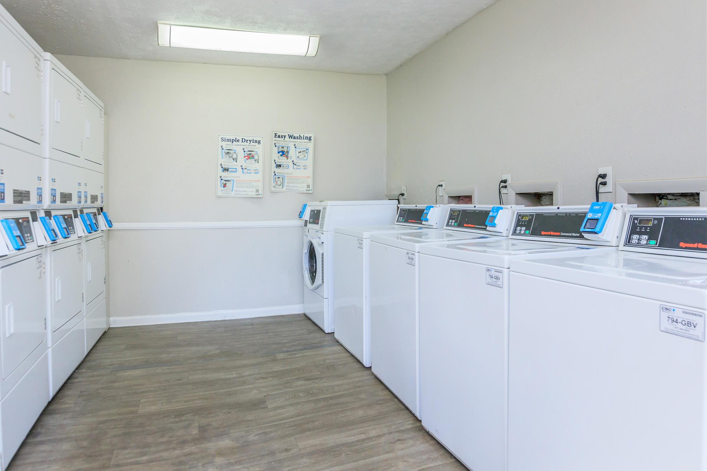A clean, well-lit laundromat featuring several white washing machines and dryers aligned against the wall. There are informational posters on the wall about laundry services. The floor has a modern wood-like finish, contributing to a tidy and organized appearance.