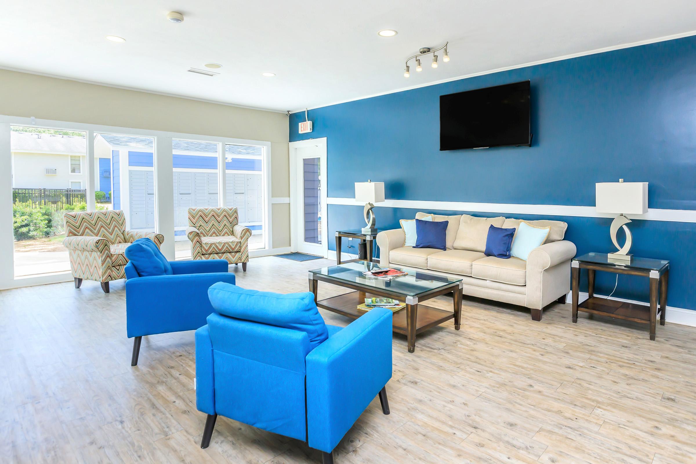 Bright and spacious lobby with blue and beige walls, featuring modern furniture including a sofa, two blue armchairs, and a glass coffee table. A flat-screen TV is mounted on the wall, and large windows allow natural light to enter. The atmosphere is welcoming and well-lit, with decorative lamps on either side of the sofa.