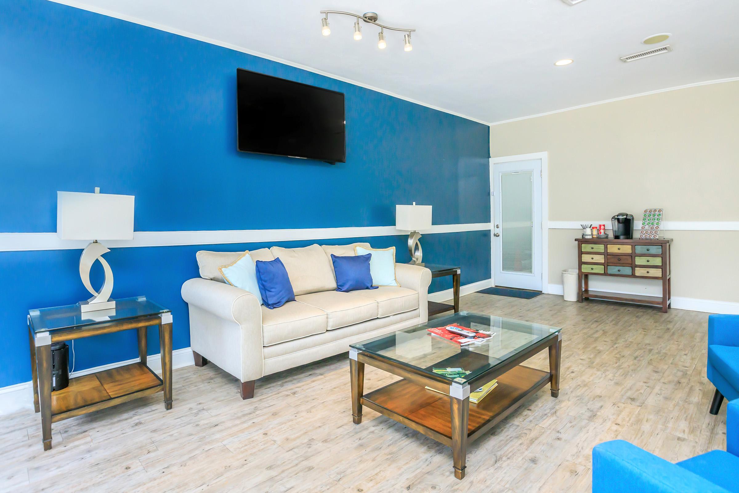 A modern lounge area featuring a beige sofa with blue cushions, two side tables with lamps, a large flat-screen TV mounted on a blue wall, and a light-colored doorway. A coffee station and a storage cabinet with drawers are visible in the background, enhancing the welcoming atmosphere.