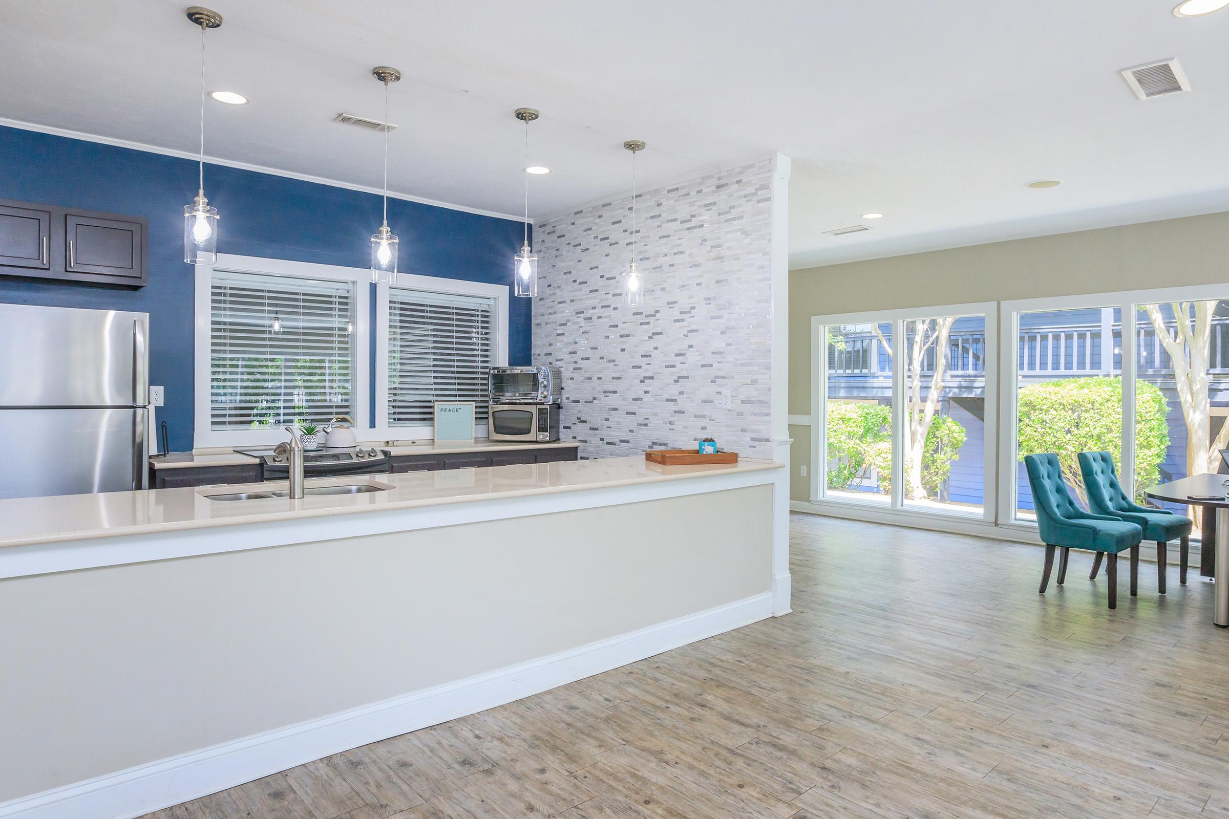 Modern open-concept kitchen featuring stainless steel appliances, quartz countertops, and pendant lighting. The kitchen flows smoothly into a bright dining area with large windows, showcasing a view of greenery outside. The floor is adorned with light-colored wood, enhancing the spacious feel of the room.