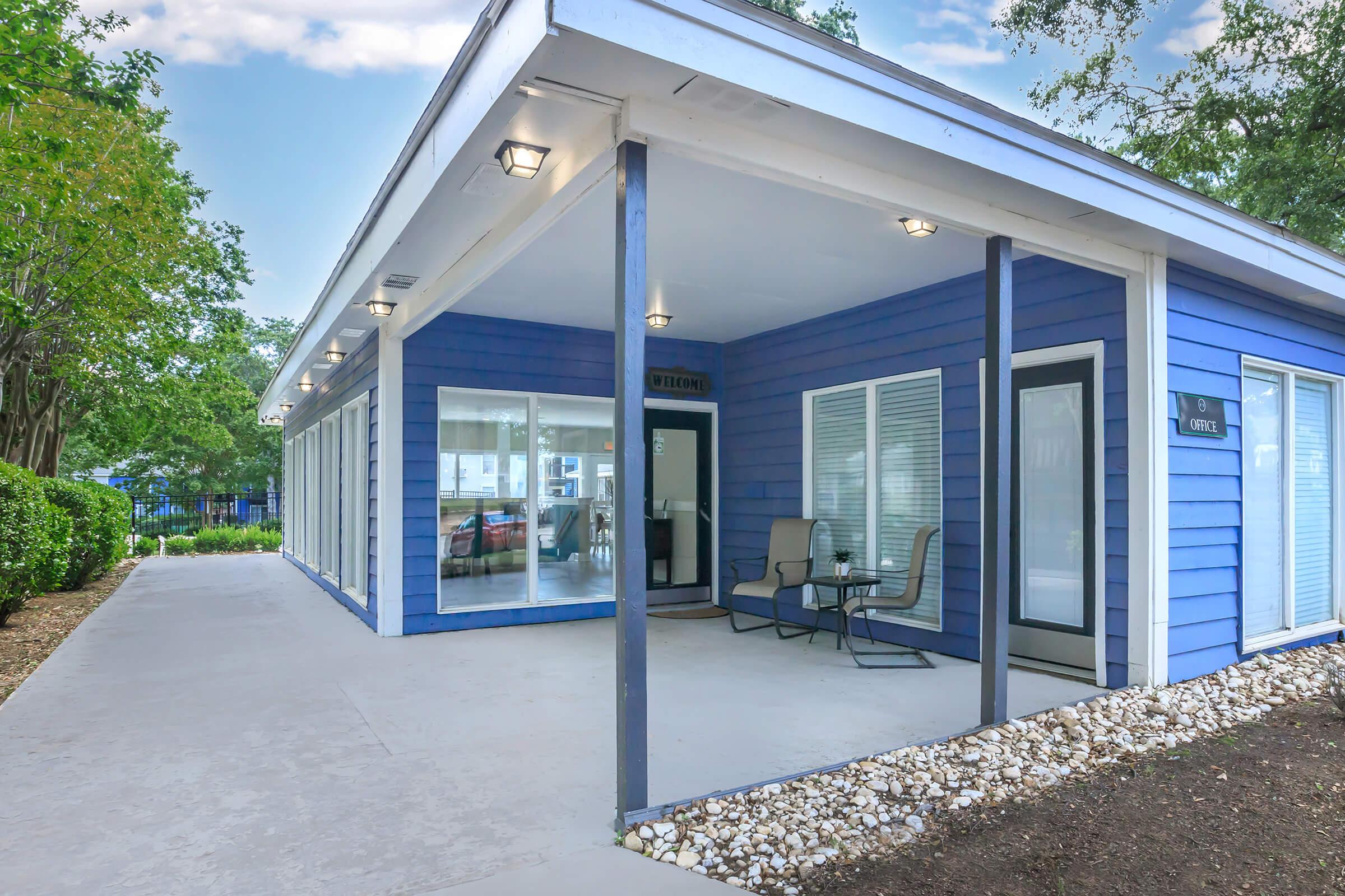 A modern blue building with large windows, featuring a covered porch and two chairs. The surroundings include green trees and a neatly landscaped area. The entrance is marked with a sign, and the overall atmosphere is inviting and serene.