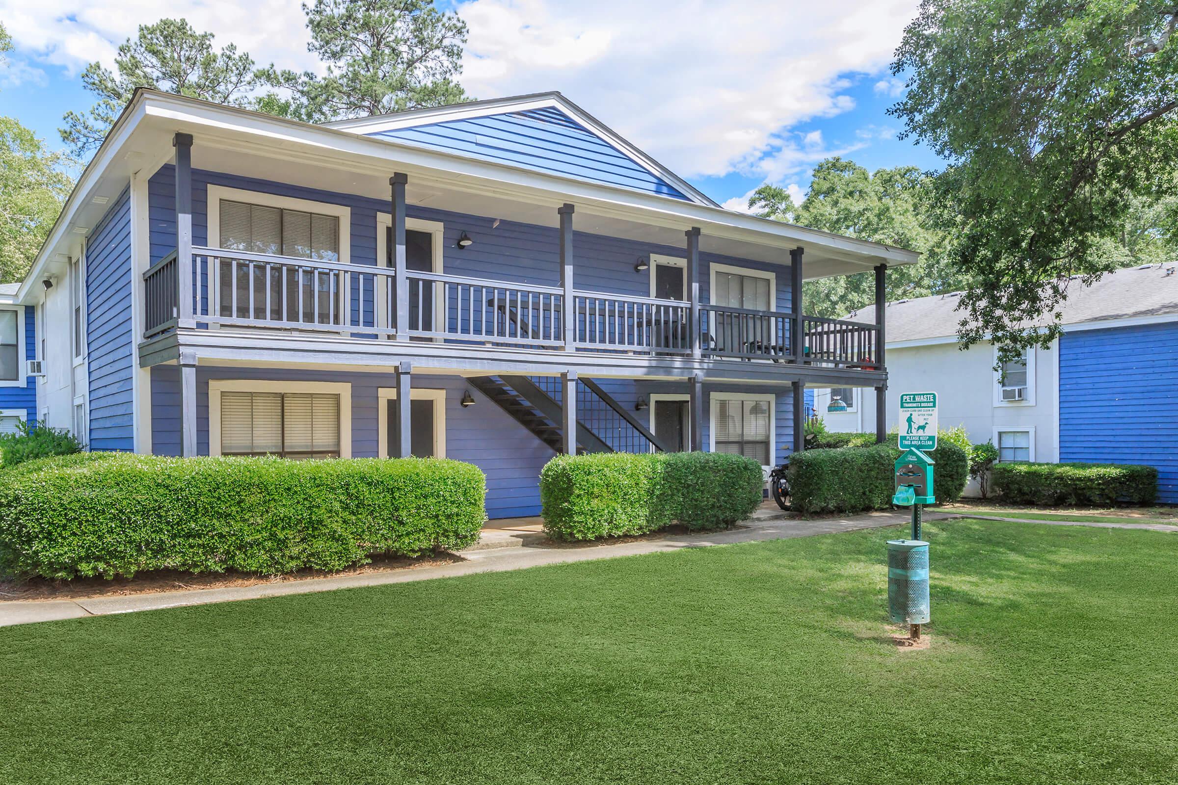 Two-story blue apartment building with a porch and balconies. Surrounded by green bushes and grass, the scene features a well-maintained landscape with signs visible nearby. The sky is partly cloudy, adding to the appealing outdoor atmosphere.